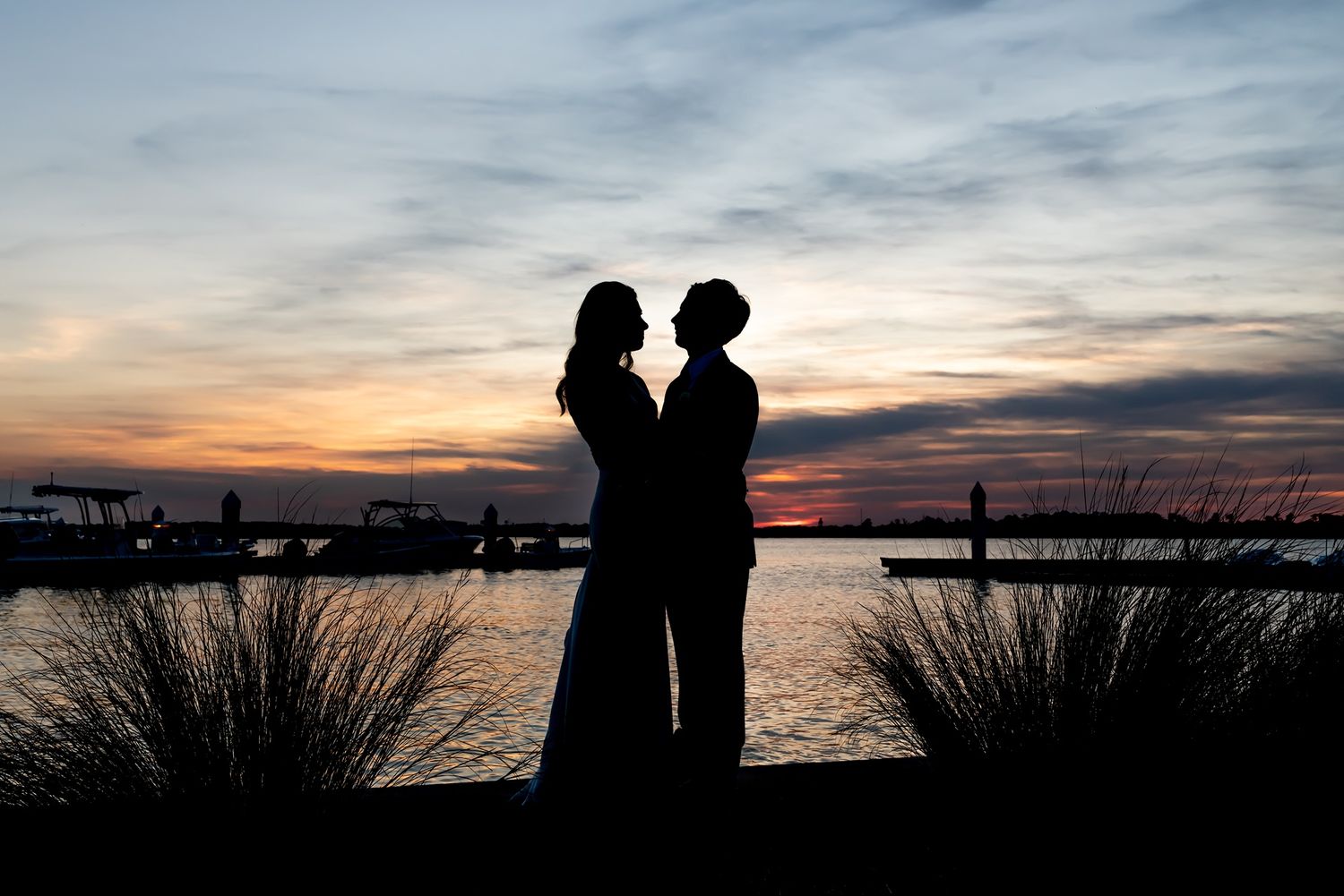 Silhouettes of couple stand together against vibrant sunset sky over waterfront with ornamental grasses in foreground.