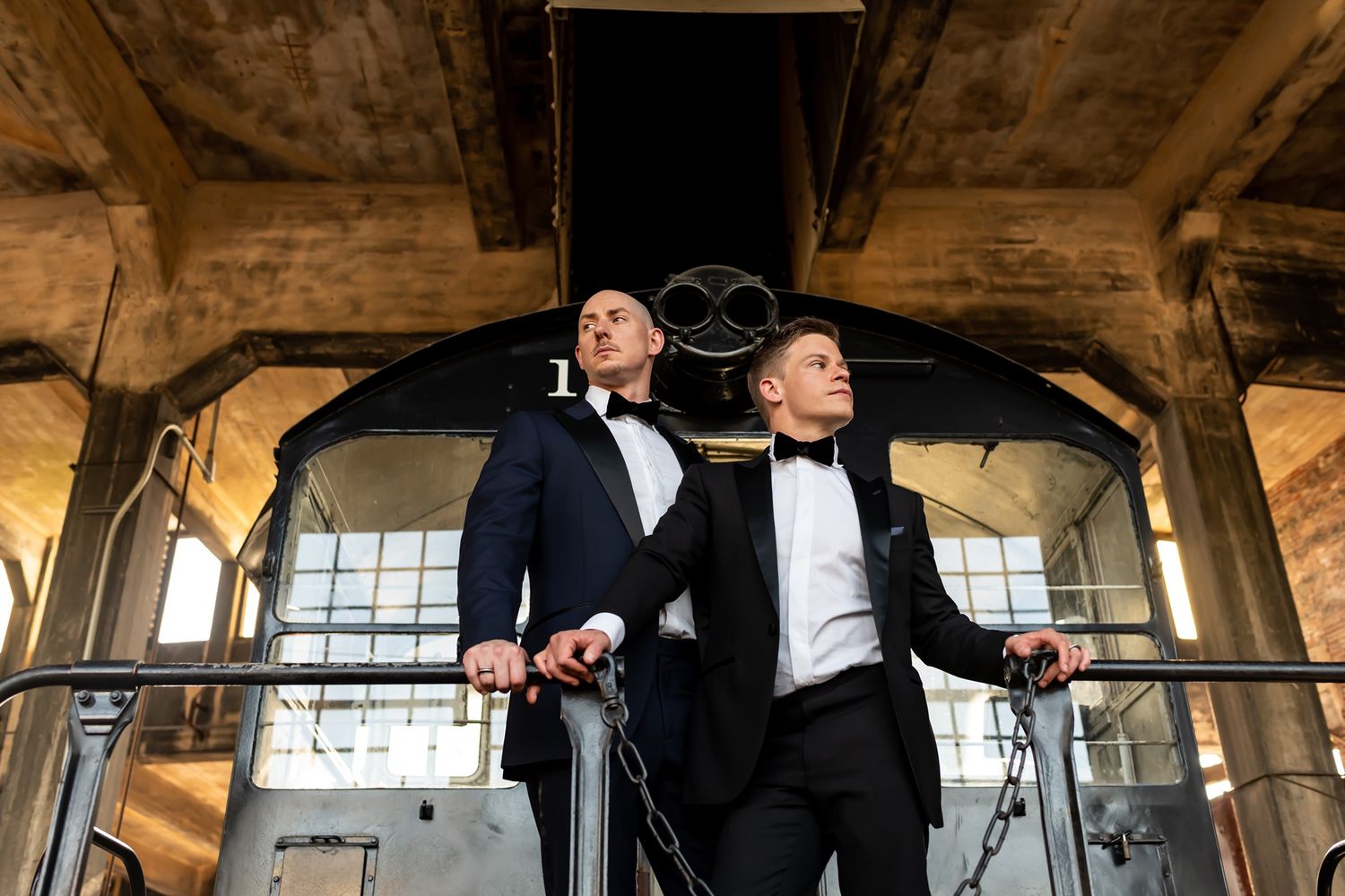 Two grooms in black tuxedos stand together on industrial staircase in rustic venue with large windows.