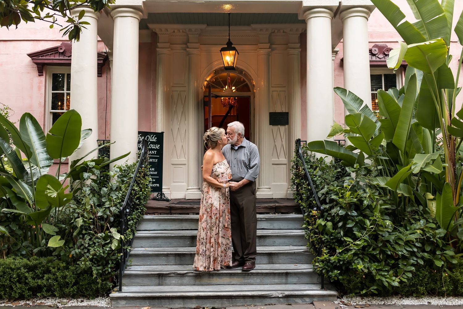 A couple stands on the front steps of a historic southern mansion with white columns and lush tropical landscaping.