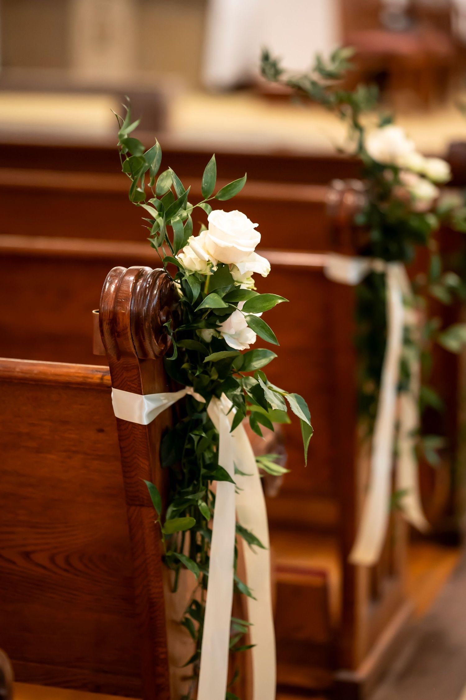 Elegant white rose and greenery floral arrangement tied with a white ribbon decorates a wooden church pew for a wedding ceremony.
