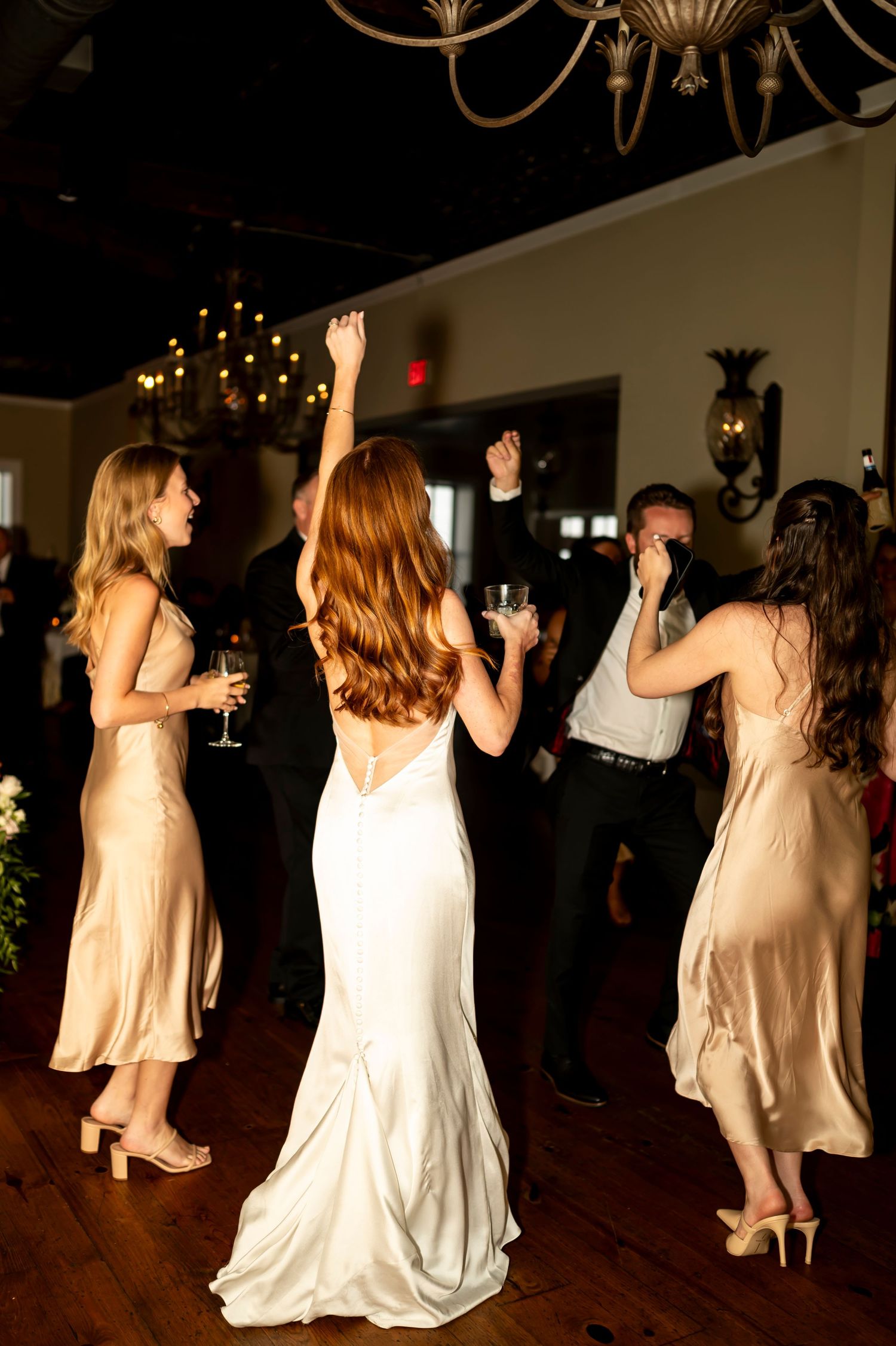 Wedding party dances on wooden floor with chandelier and string lights overhead, bride in white gown surrounded by bridesmaids.