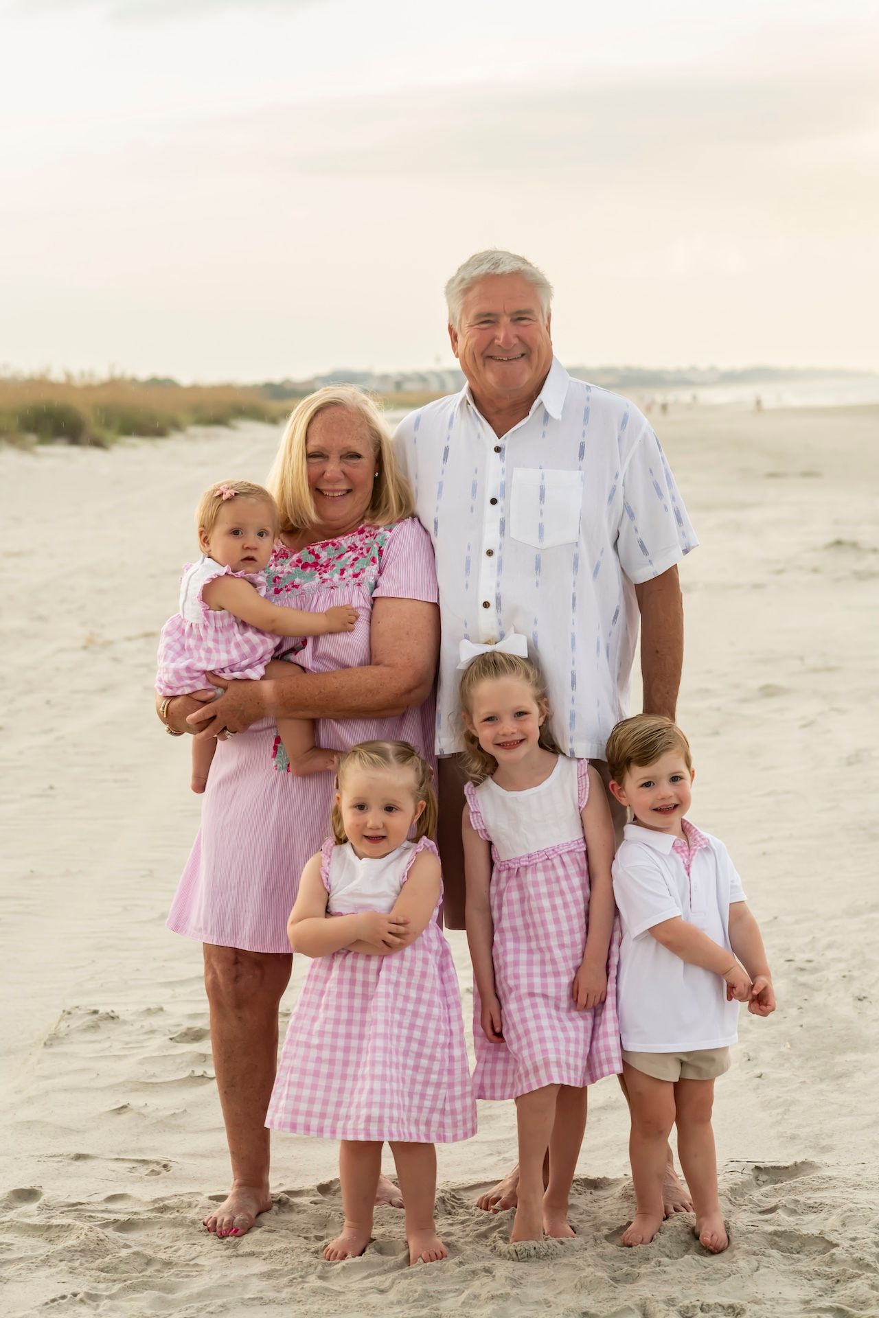 Family group wearing coordinated pink and white outfits poses together on sandy beach at sunset.