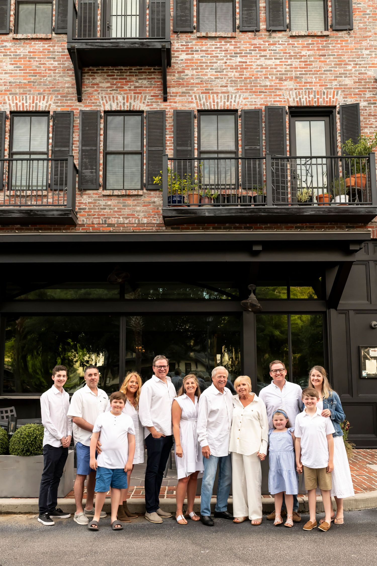 Family group in white shirts poses together in front of a brick building with ornate balconies and window shutters.