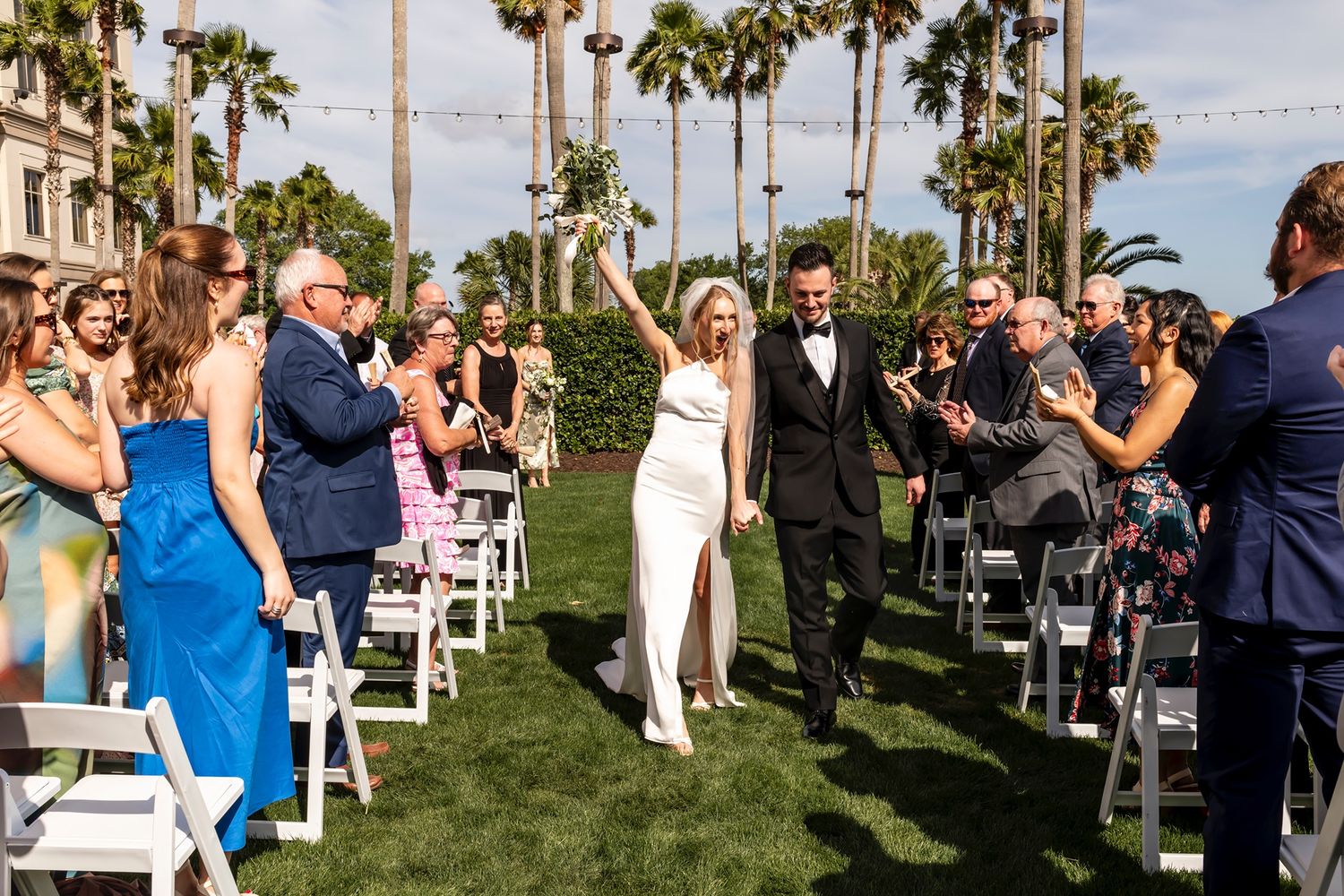Newly married couple exits down outdoor aisle lined with palm trees and happy guests during sunny wedding ceremony.