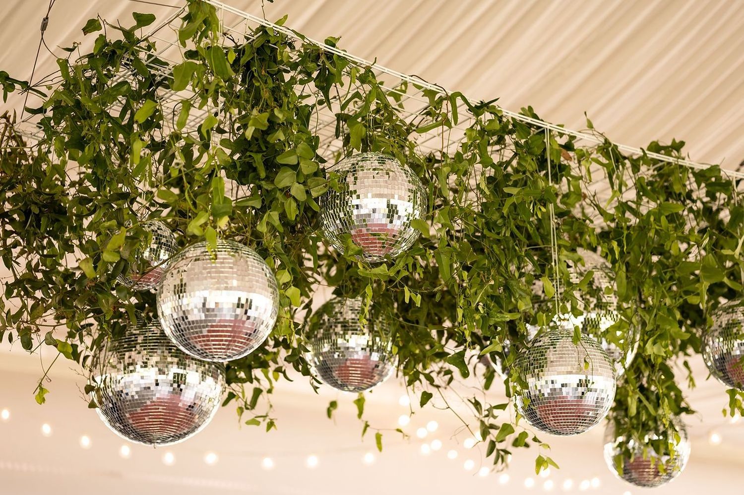 Mirrored disco balls hang from the ceiling amongst green ivy vines and twinkling string lights at an event venue.