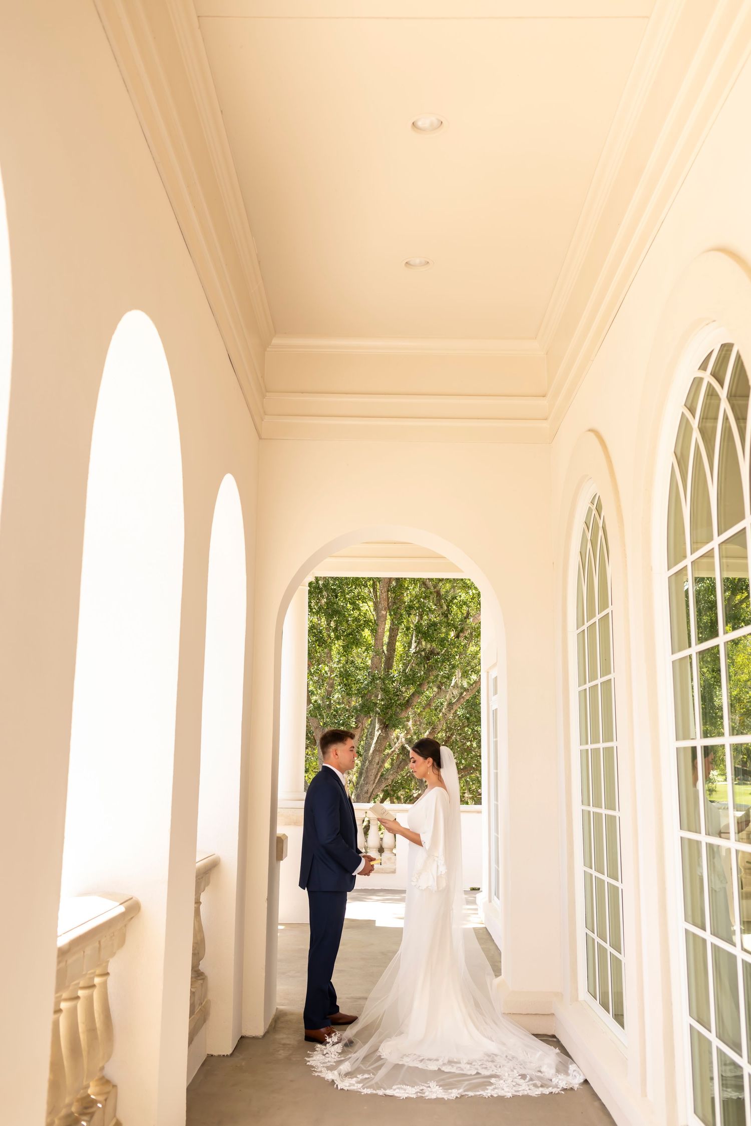 Elegant couple stands in a bright white arched colonnade with large windows and lush greenery visible outside.