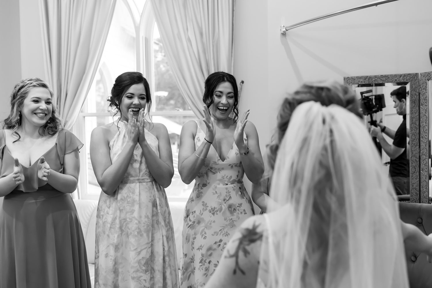 A group of bridesmaids in a boutique dressing room react joyfully while looking at someone in a wedding dress.