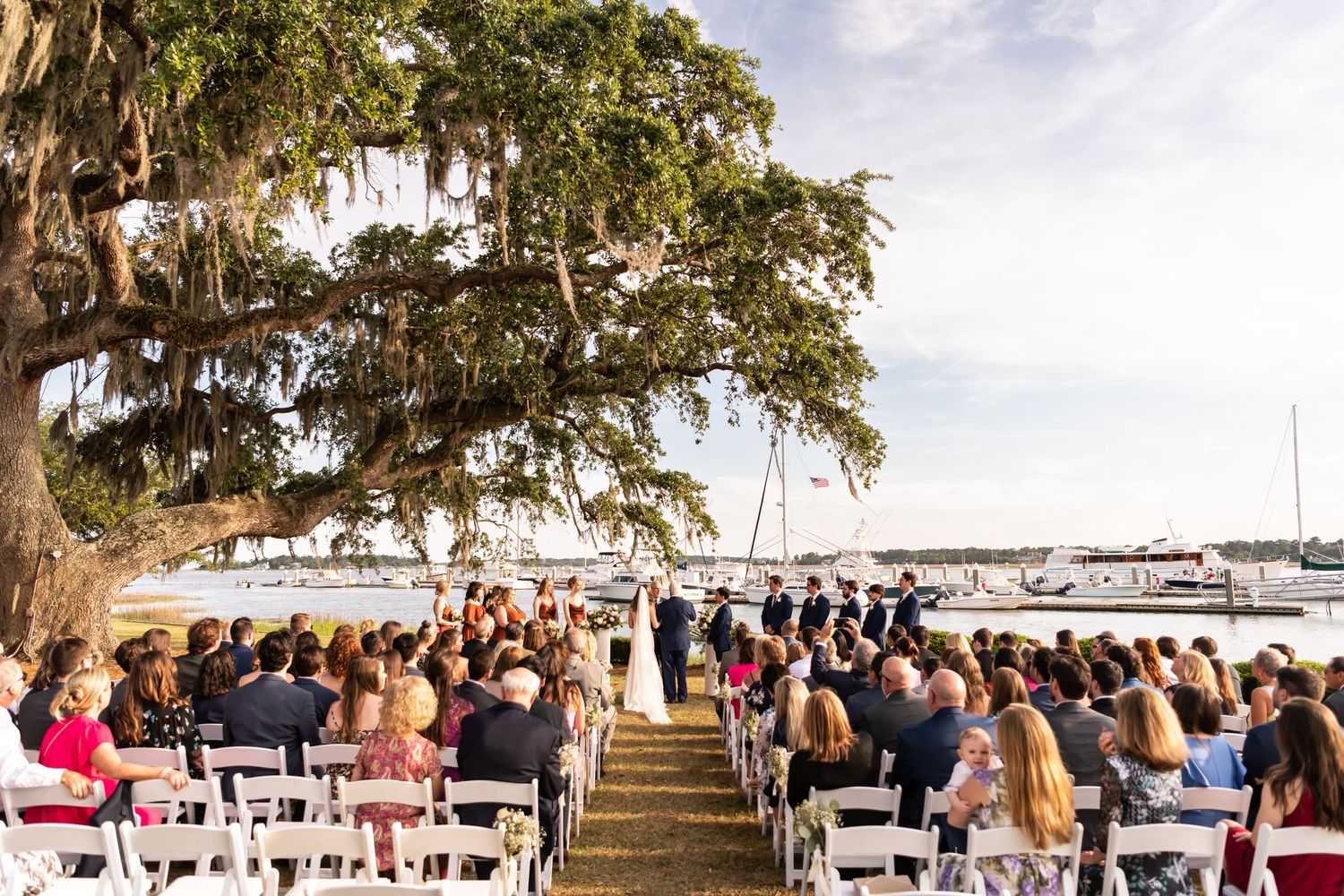 Wedding guests gather under Spanish moss-draped oak trees near a waterfront dock at an outdoor ceremony.