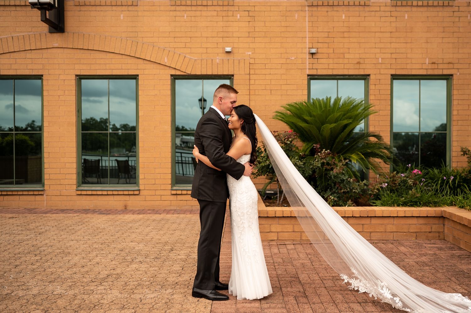 A couple in formal wedding attire shares a romantic moment outside a modern brick building with glass windows.