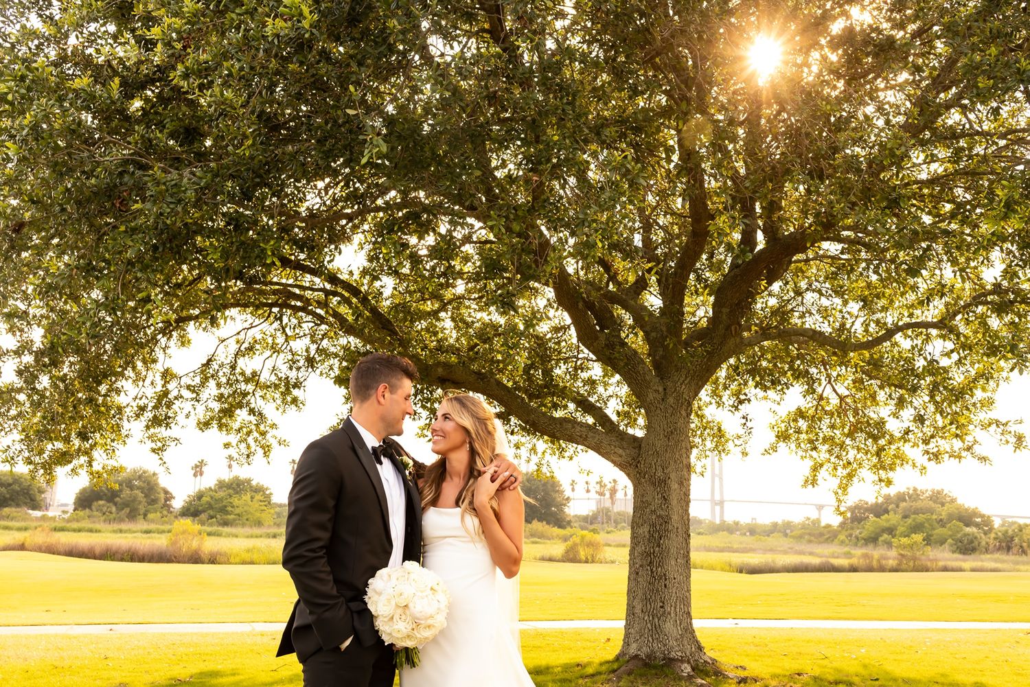 A couple poses beneath a majestic oak tree during golden hour sunset on a grassy field.