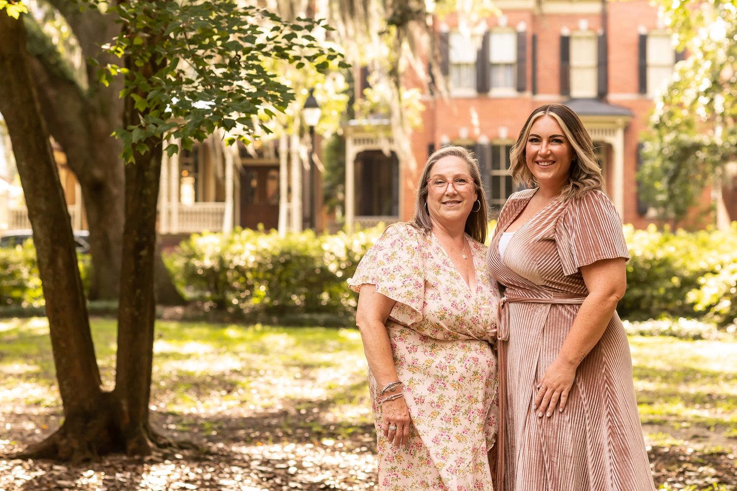 Two people wearing floral and mauve dresses pose together in a sunlit garden setting with brick buildings in background.