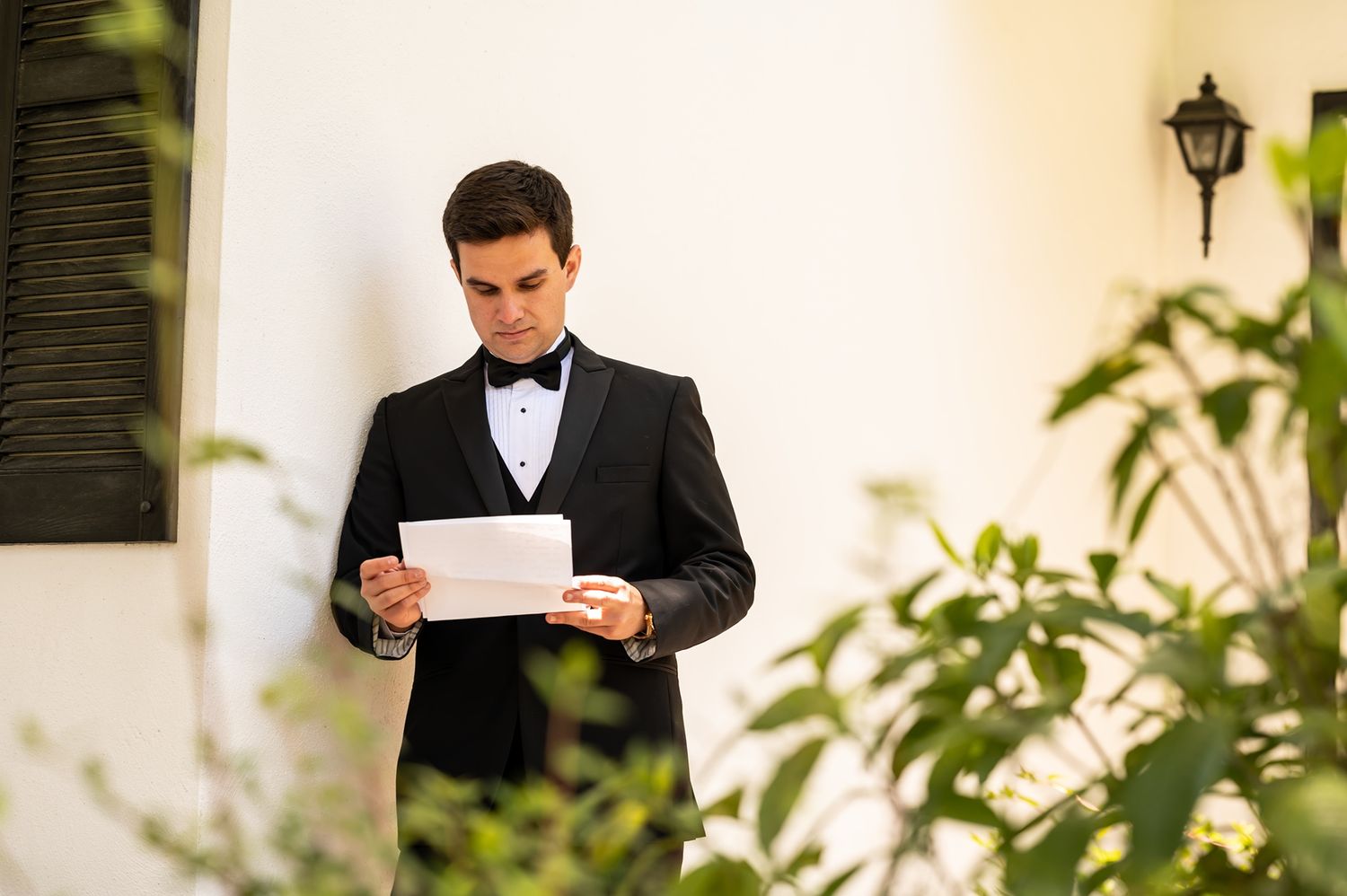 A person in a black tuxedo and bow tie stands outdoors reading papers near greenery and a white wall.