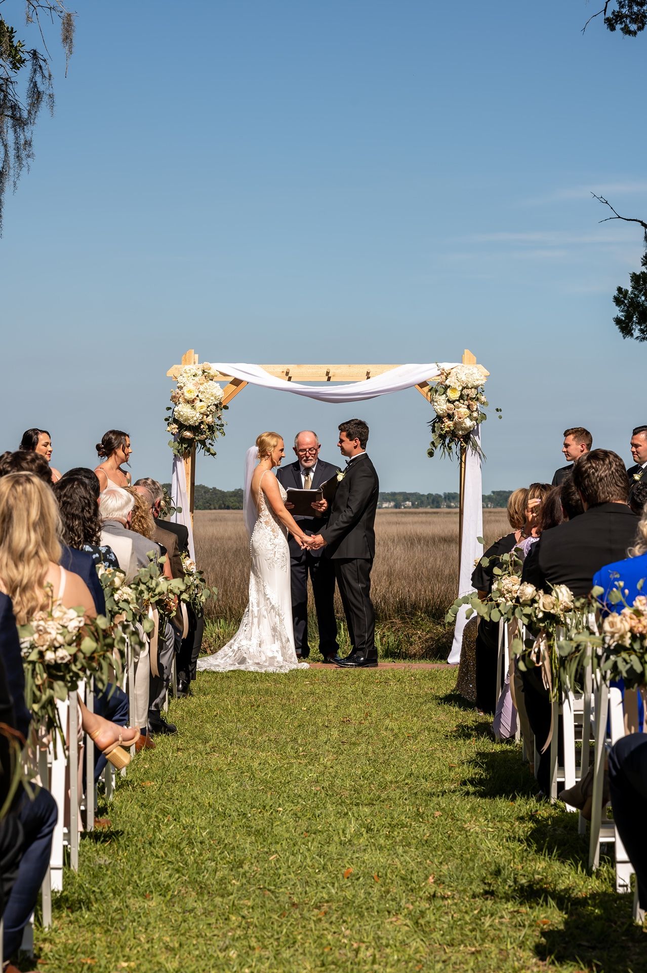 Outdoor wedding ceremony on grassy lawn overlooking marsh with white floral arch and rows of white chairs.