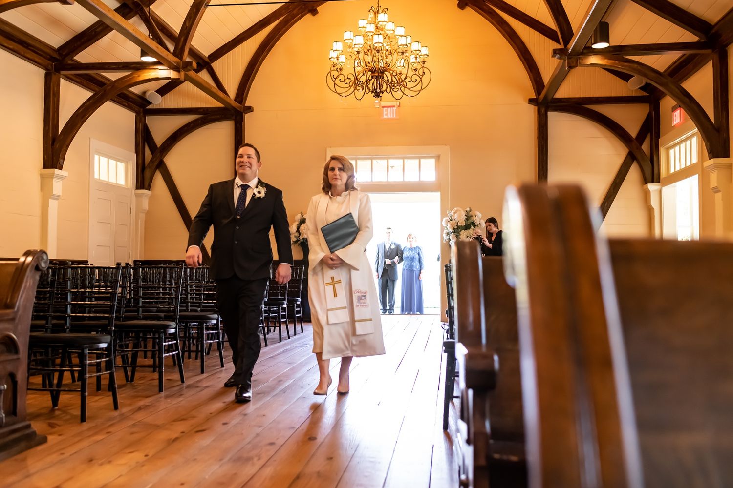 People walking down a wooden aisle in an elegant chapel venue with exposed beams and a crystal chandelier overhead.
