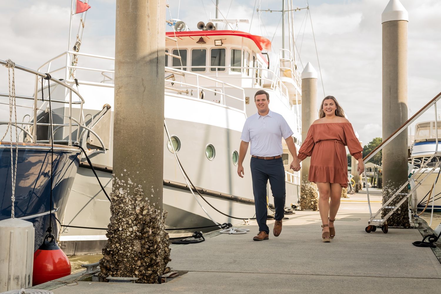 A couple walks hand in hand along a marina dock with boats and vessels moored in the background on a sunny day.