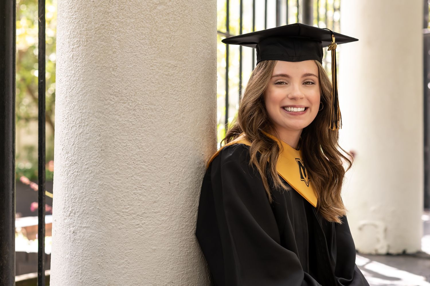 Graduate in black cap and gown and yellow shirt smiles while leaning against white column.
