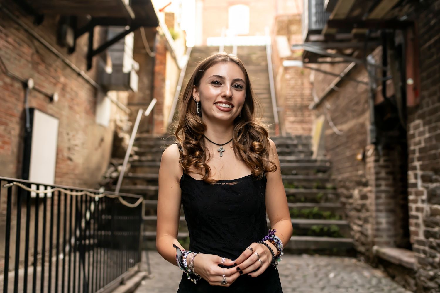 Woman in black dress standing in urban alleyway with brick walls and stairs behind her in warm lighting.