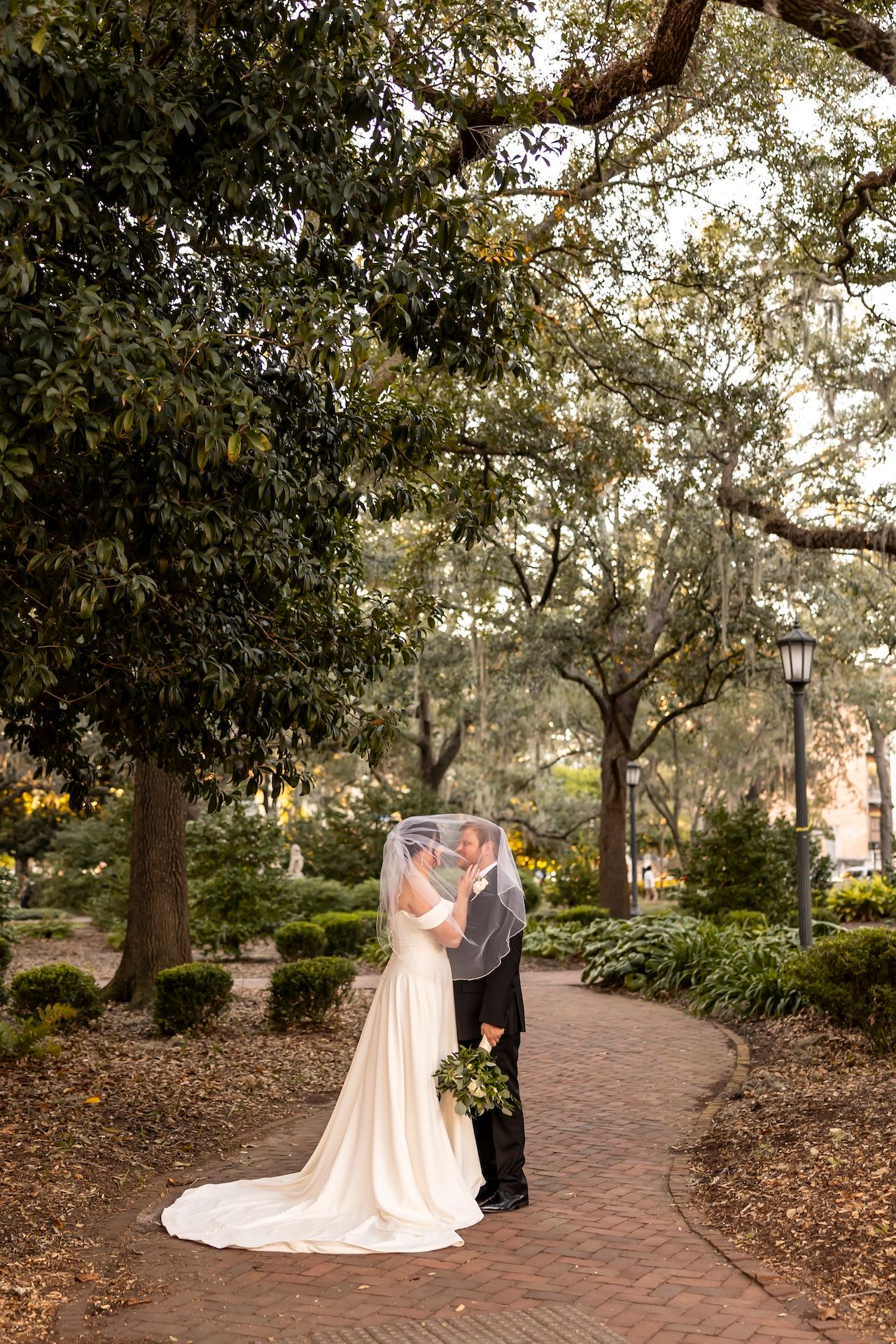 Two people in wedding attire stand lovingly on a brick path surrounded by trees in a romantic garden setting.