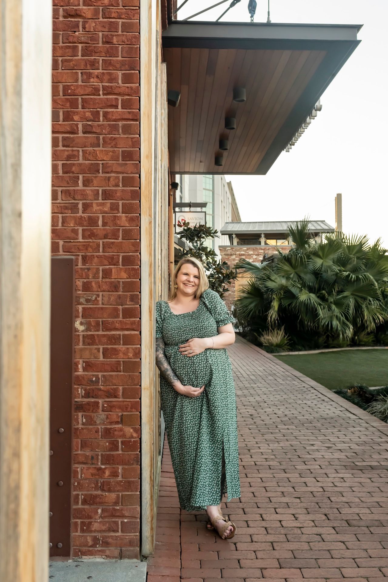 A person in a green polka dot dress poses on brick steps in an outdoor setting with brick walls and palm plants visible.