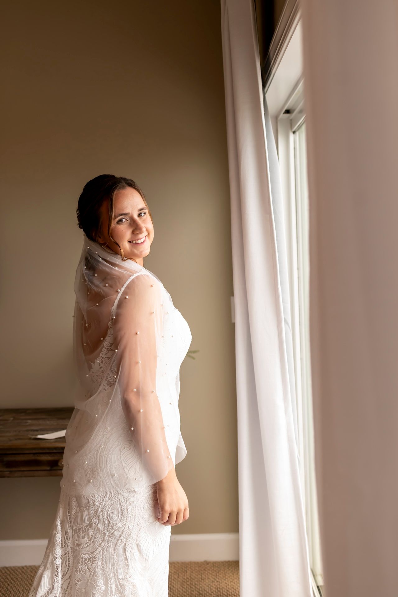 Bride in elegant white dress and veil poses in different angles by a window with natural light streaming in.