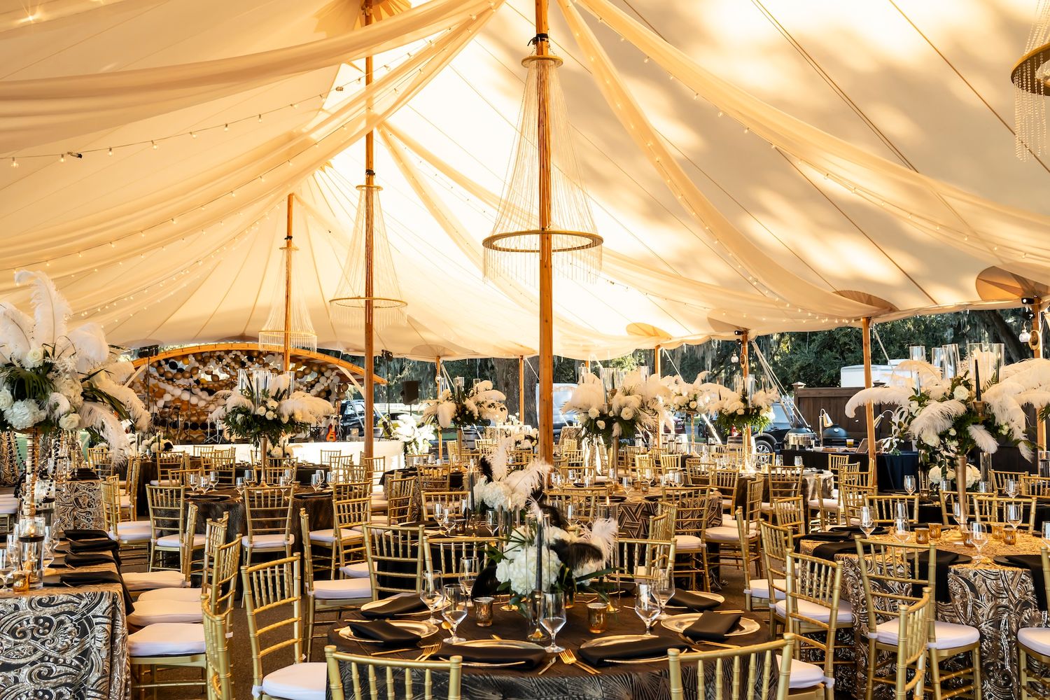 An elegant wedding reception tent decorated with warm lighting, gold chairs, and floral centerpieces for an evening celebration.