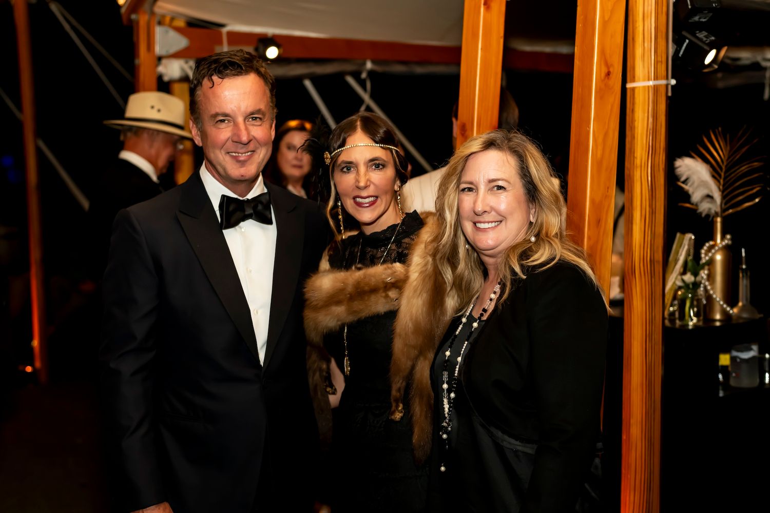 Three people in formal evening attire smile while attending an elegant event.