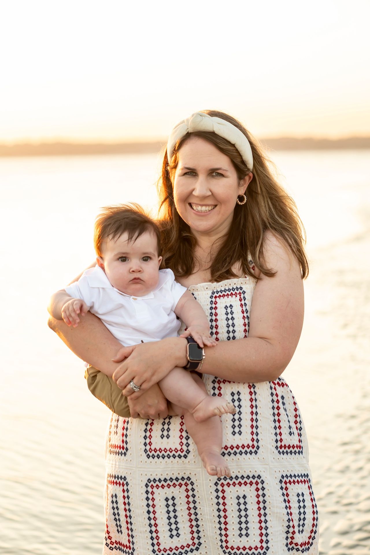 A parent and baby stand together on a beach at sunset, wearing summer attire with a geometric patterned dress.