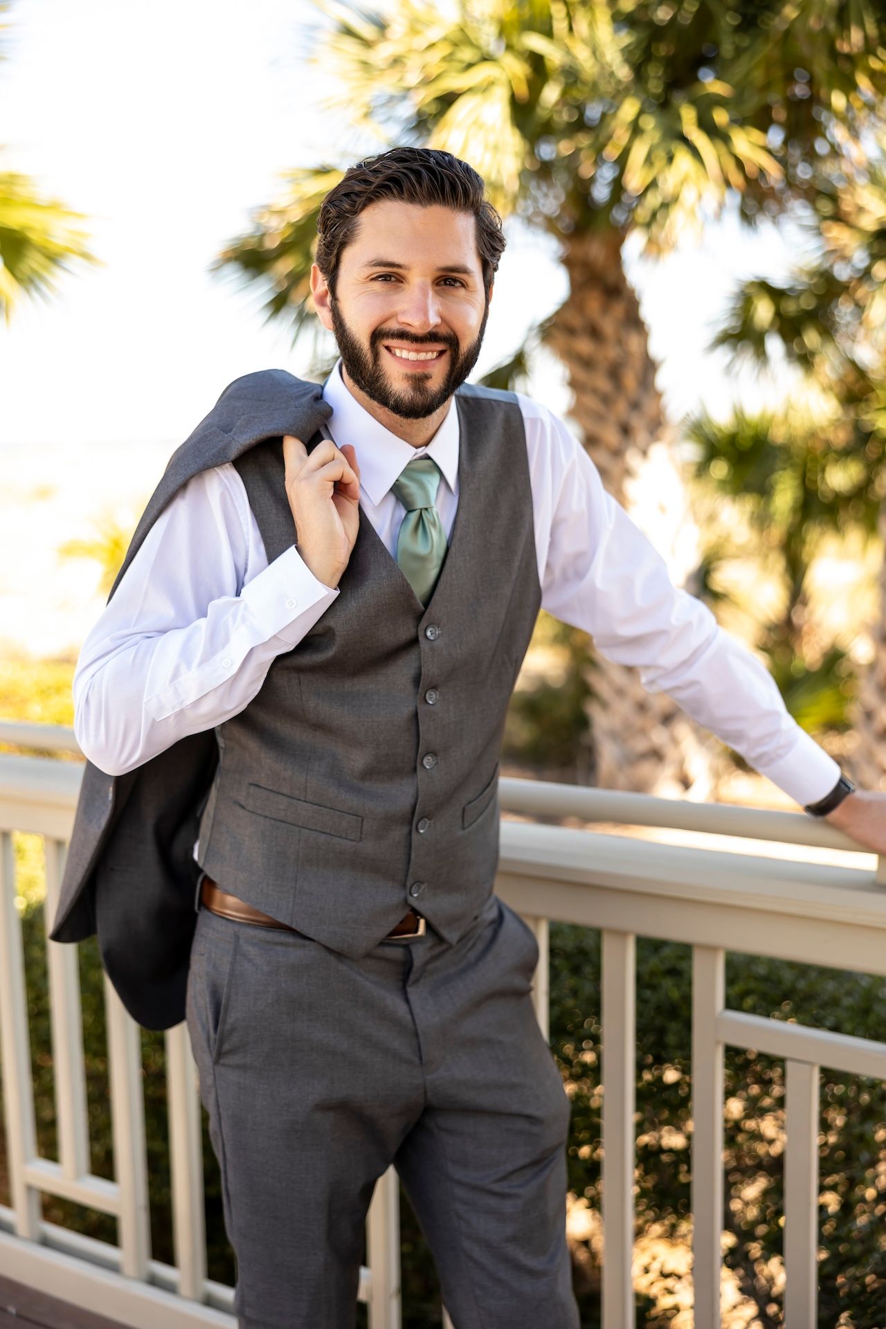 Well-dressed person in a gray vest and tie posing outdoors against a palm tree backdrop on a sunny day.