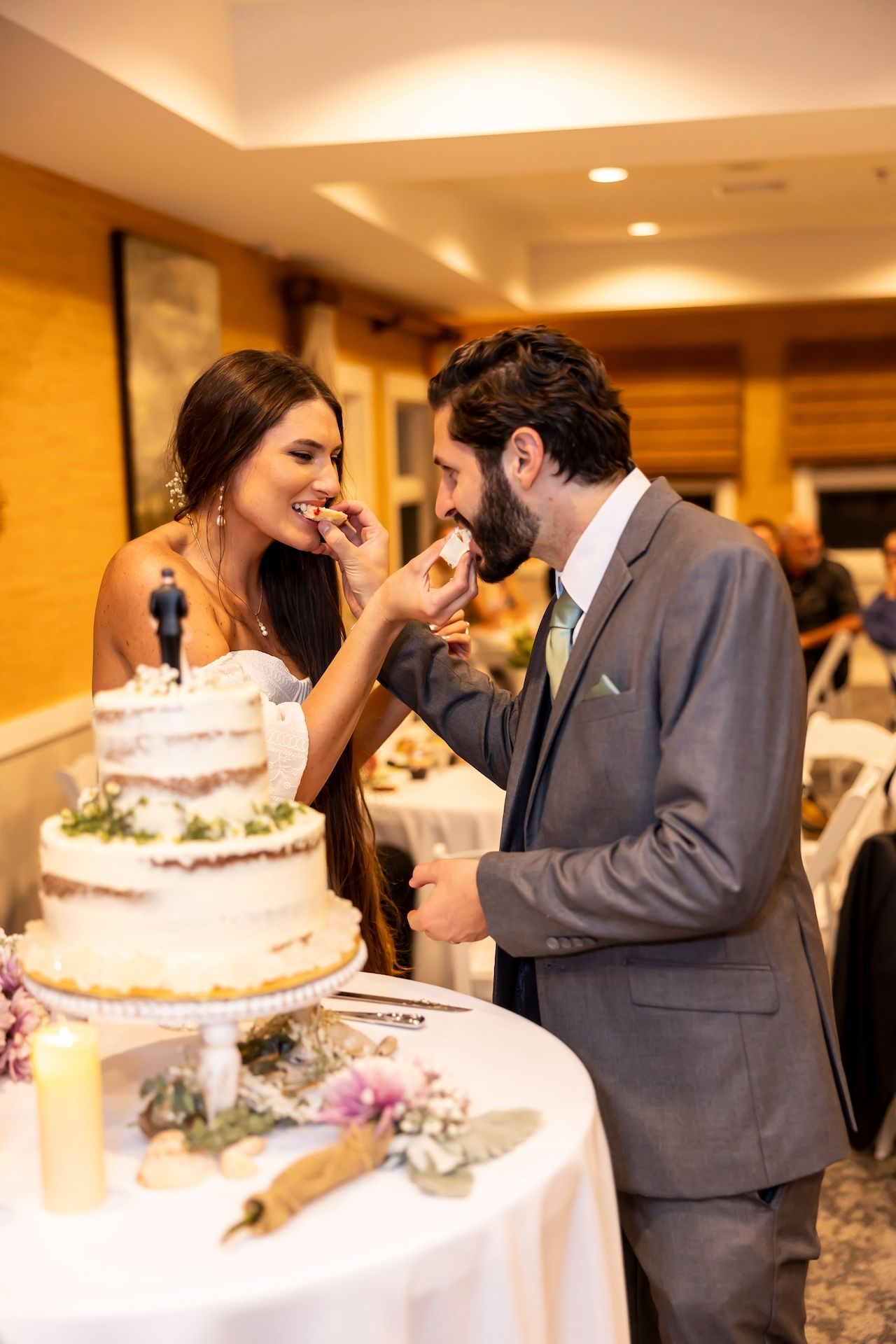 A couple shares their wedding cake at a reception in a warmly lit banquet room, feeding each other with joyful expressions.