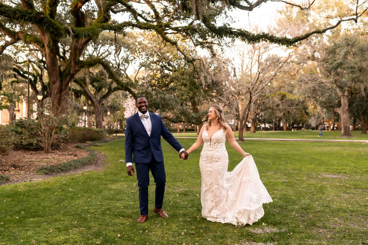 A couple walks hand in hand through a picturesque garden path lined with trees at sunset.