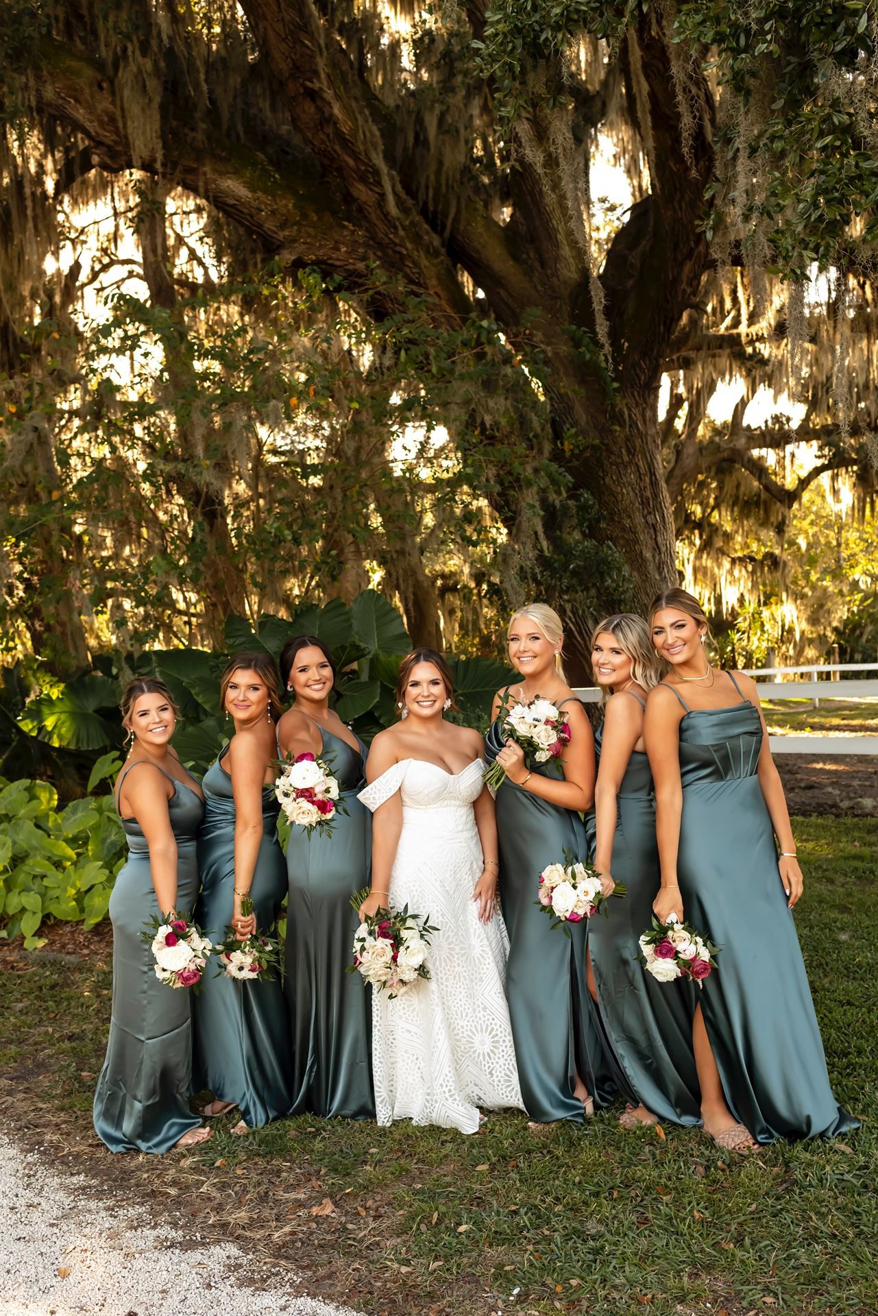 Wedding party in dusty blue bridesmaid dresses poses with bride under Spanish moss draped trees in outdoor setting.