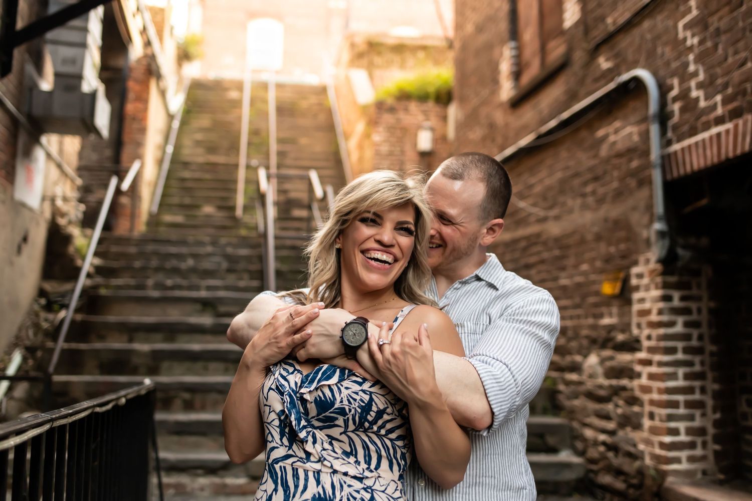 A joyful couple laughs together while embracing on outdoor brick stairs during a romantic evening photoshoot.