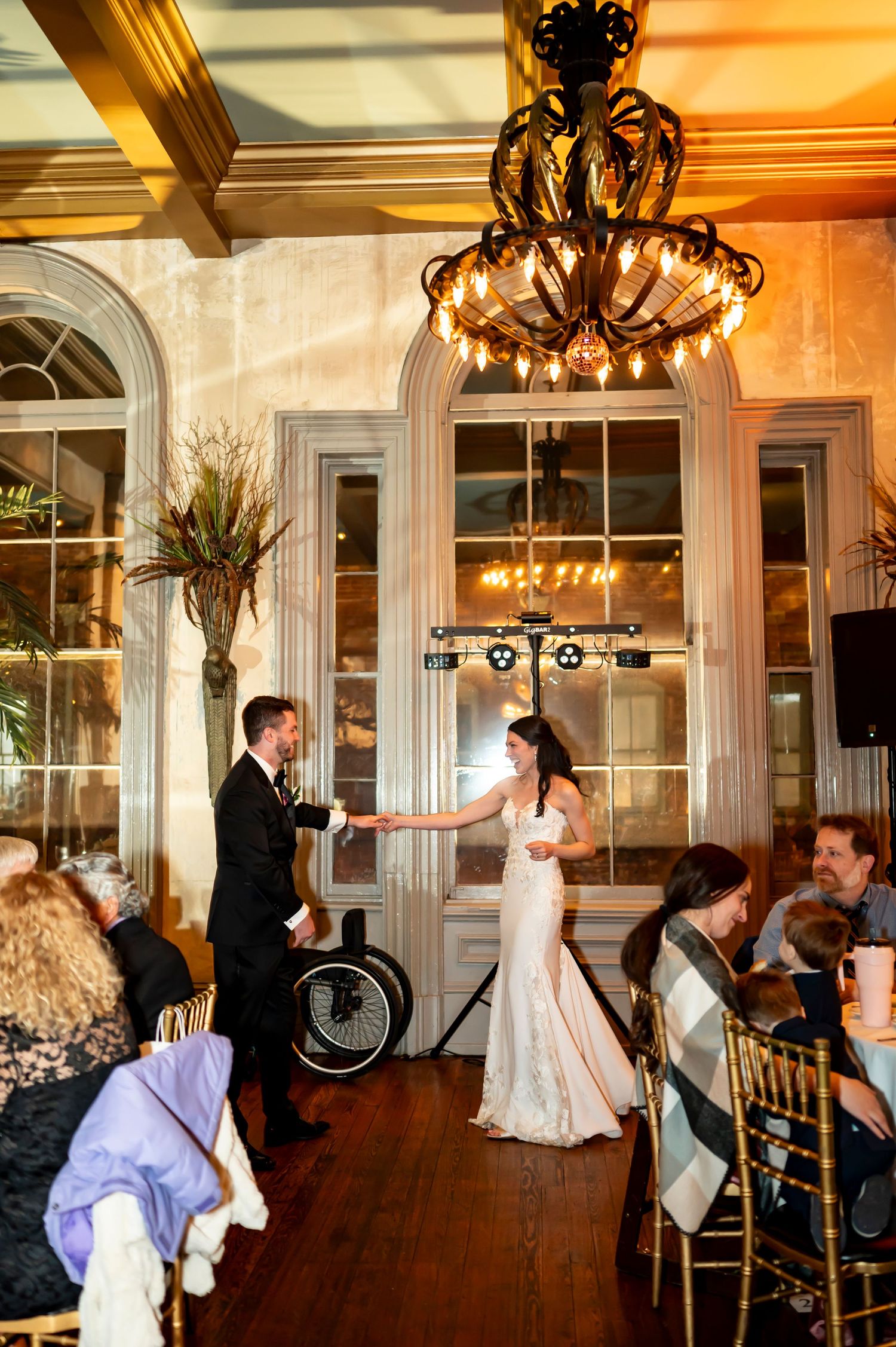 Two people dance elegantly in a grand ballroom under an ornate chandelier with guests seated at tables along the sides.