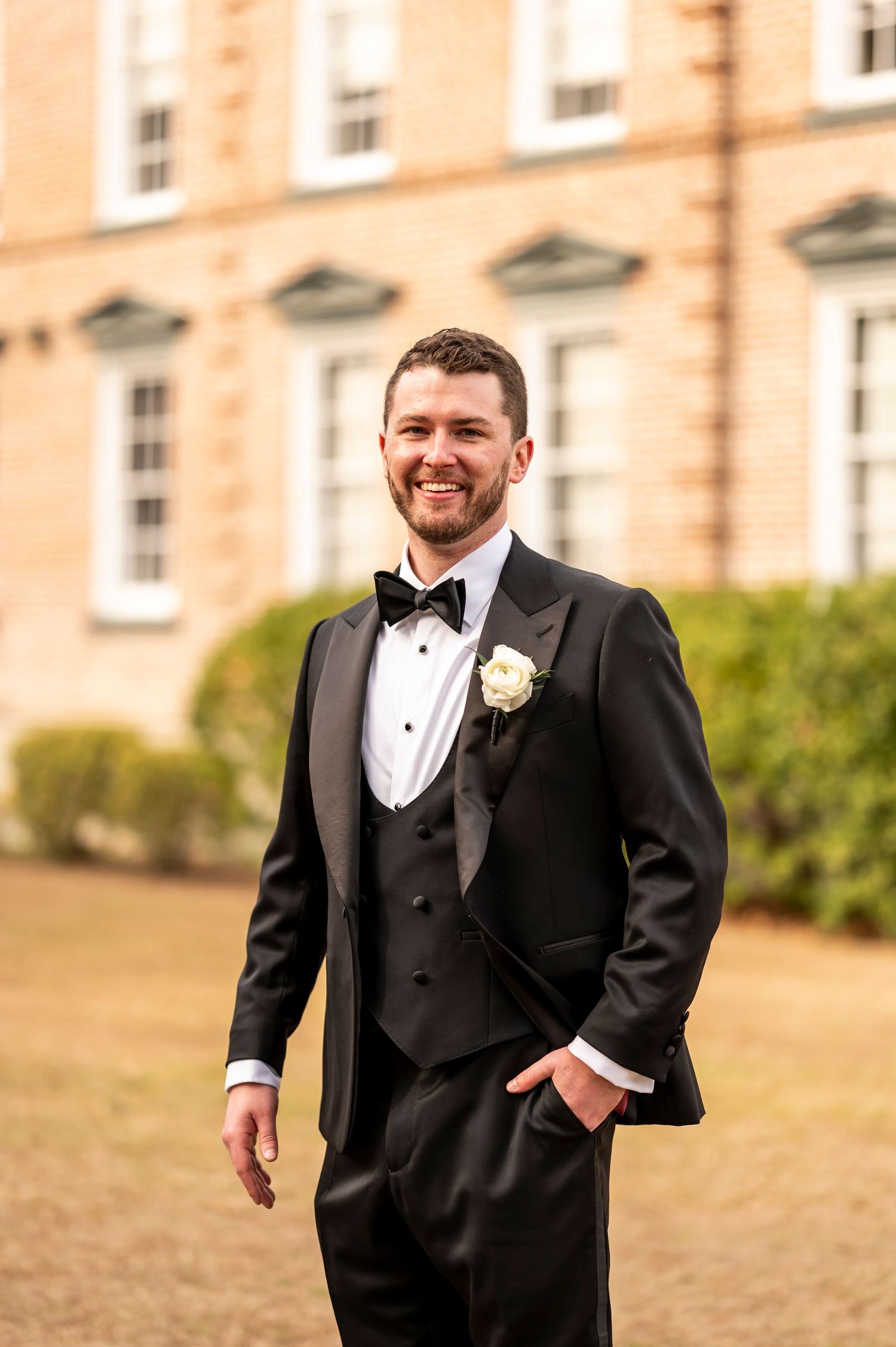 A groom in a classic black tuxedo with bow tie and boutonniere poses outdoors in front of a brick building.