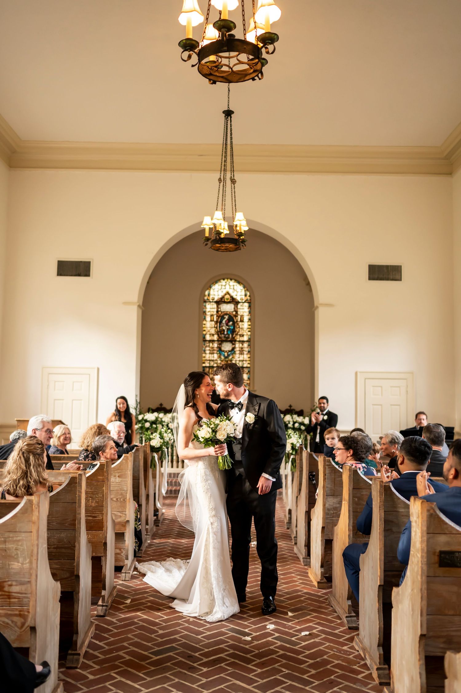 A couple walks down the aisle of a historic church with brick floors and elegant chandeliers as guests look on from wooden pews.