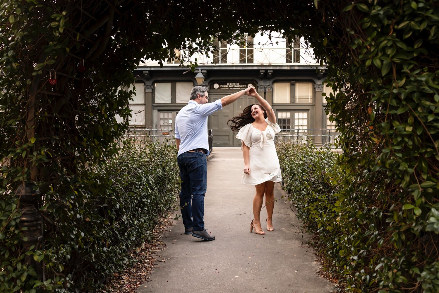 Two people dancing together joyfully on a garden path framed by green hedges with a building in the background.