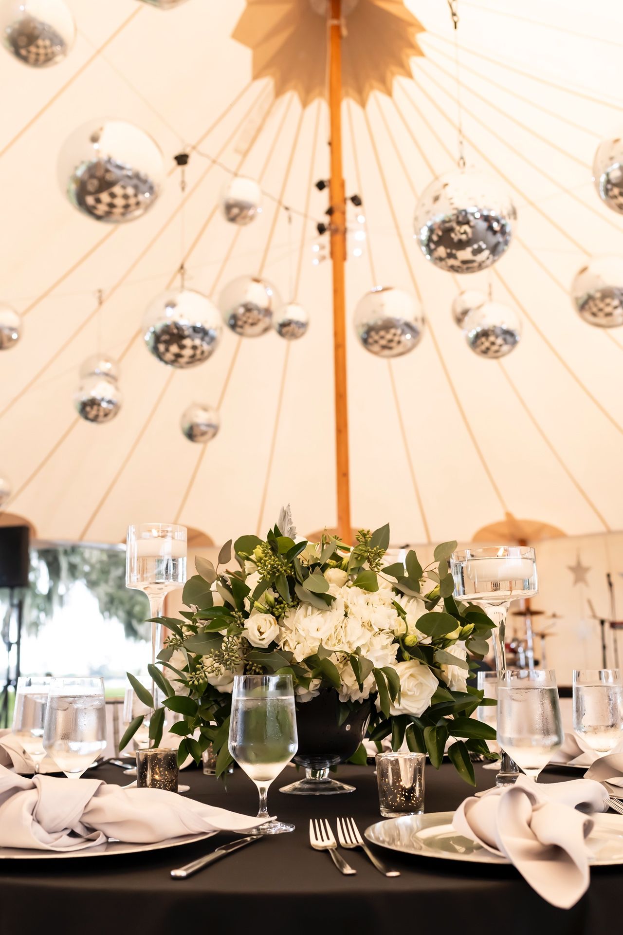 White floral centerpiece on dark tablecloth under cream canvas tent with disco balls and orange pole at wedding reception.