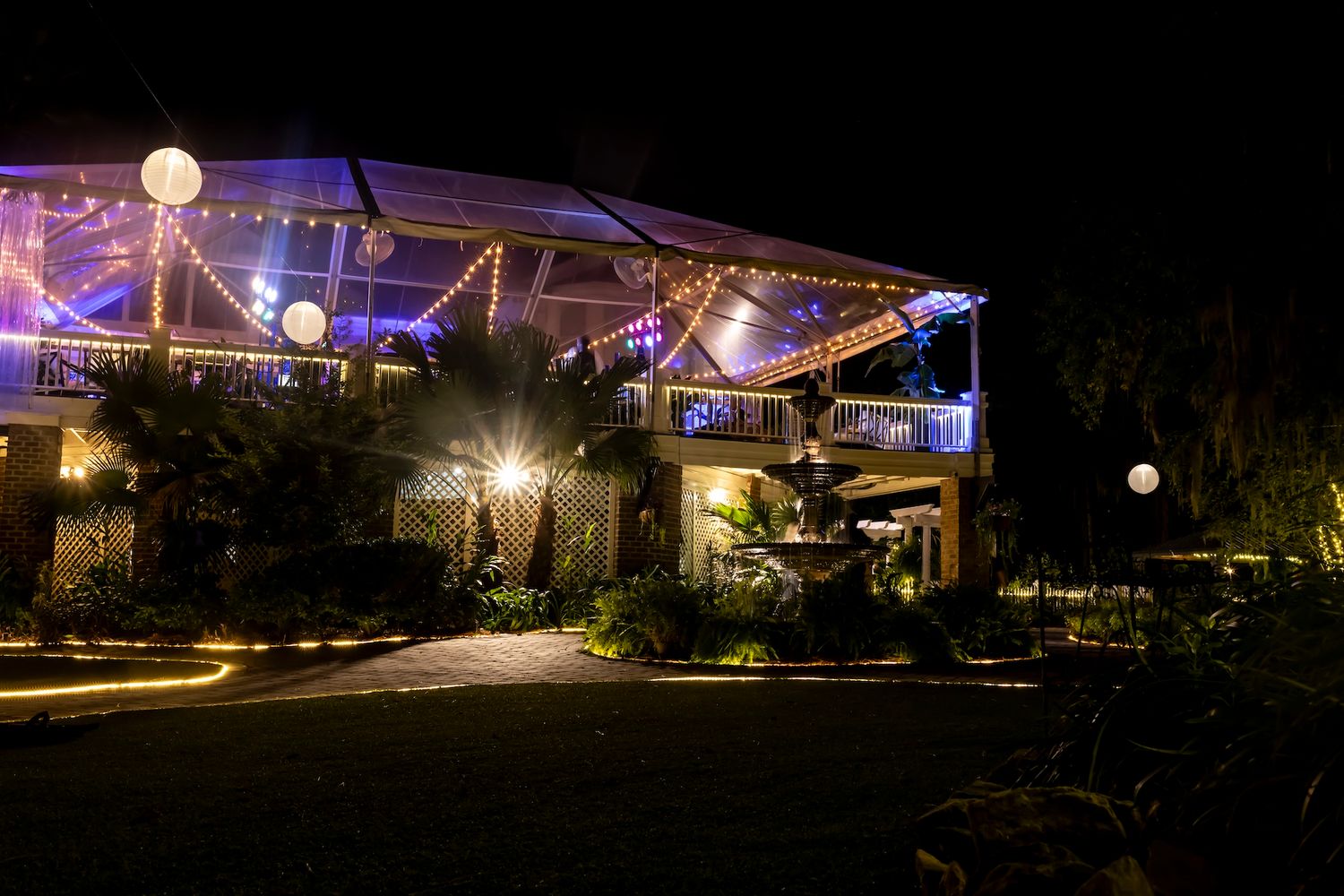 An illuminated outdoor event venue with string lights and purple uplighting creating a romantic nighttime atmosphere.