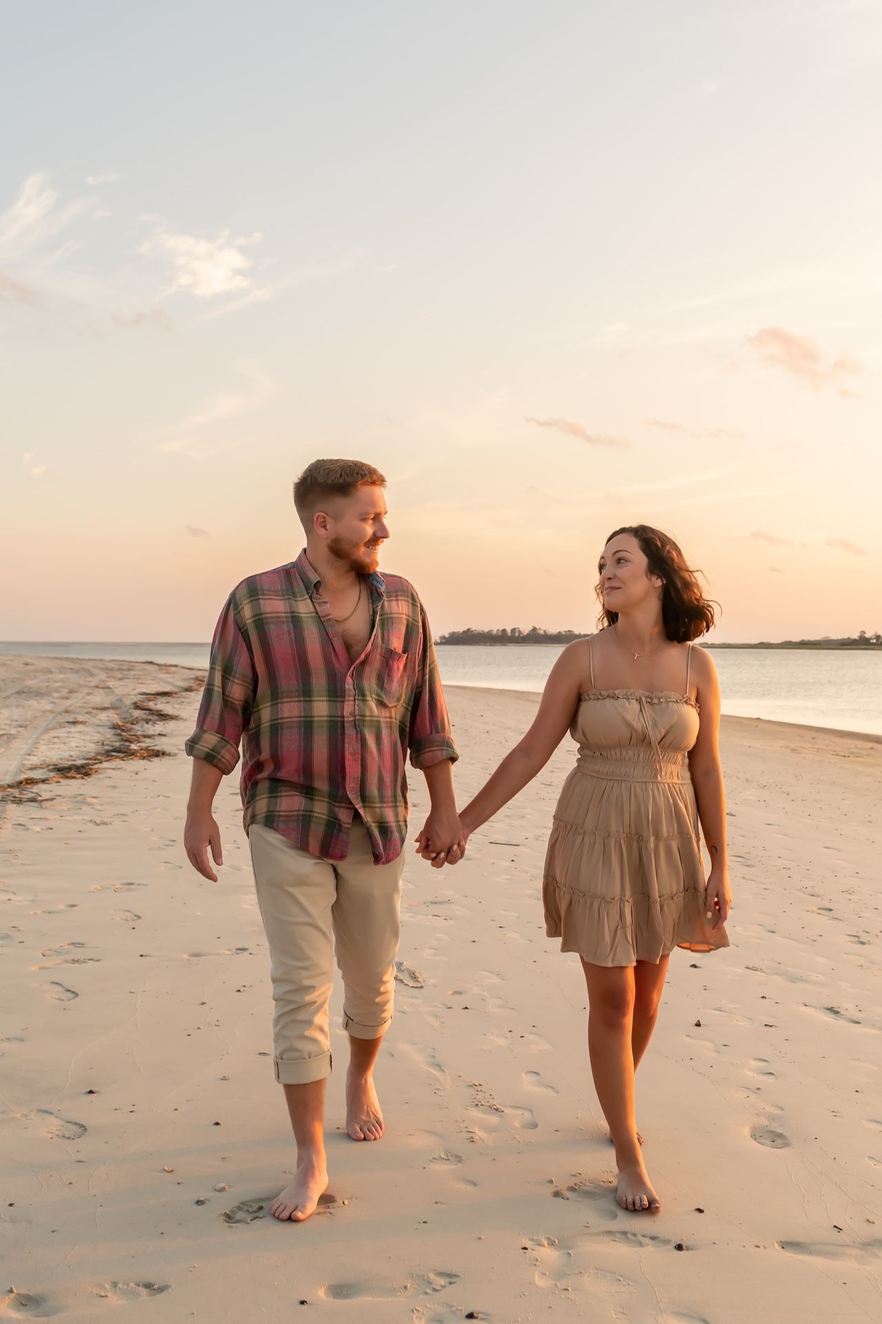 A couple walks hand in hand along a sandy beach at sunset, with waves and shore in the background.