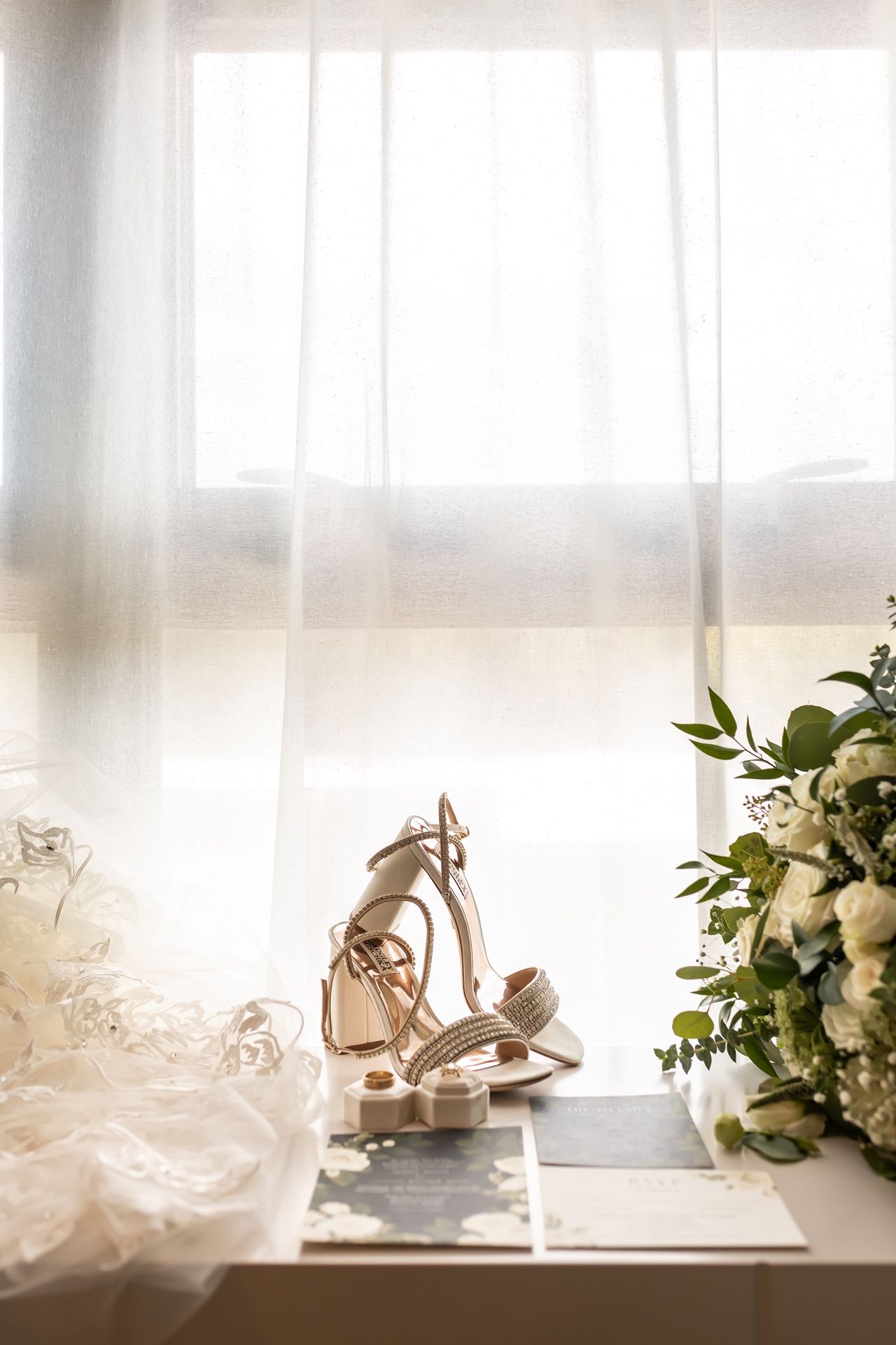 Elegant white wedding heels and bouquet arranged by sunlit window with sheer curtains casting soft light.