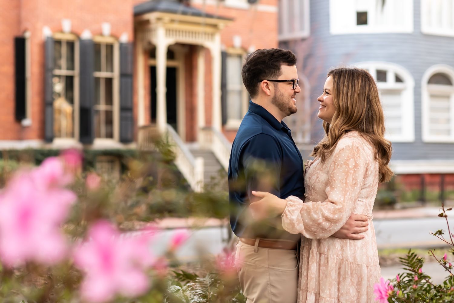 Two people embrace romantically in front of historic buildings with pink flowers blooming in the foreground.