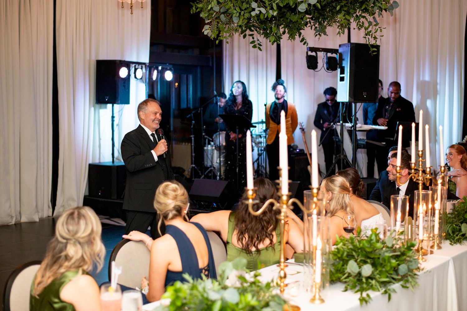 Wedding guests seated at an elegant reception table with lit candles while a speaker addresses the crowd from a podium.