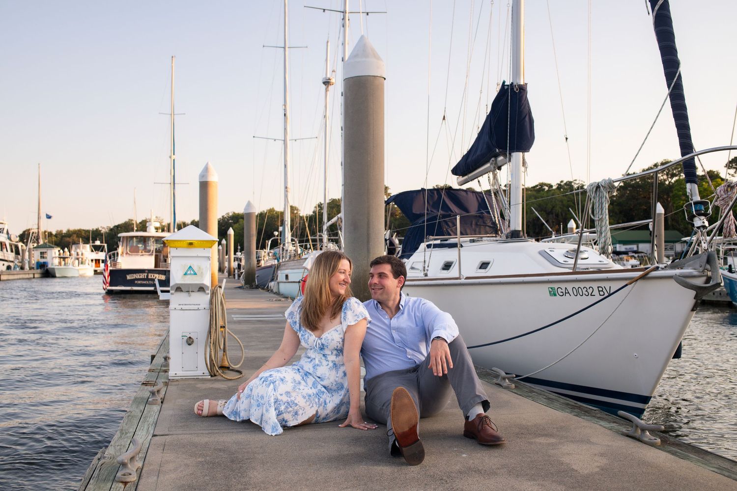 A couple sits together on a dock at Isle of Hope marina with sailboats and yachts in the background at sunset.