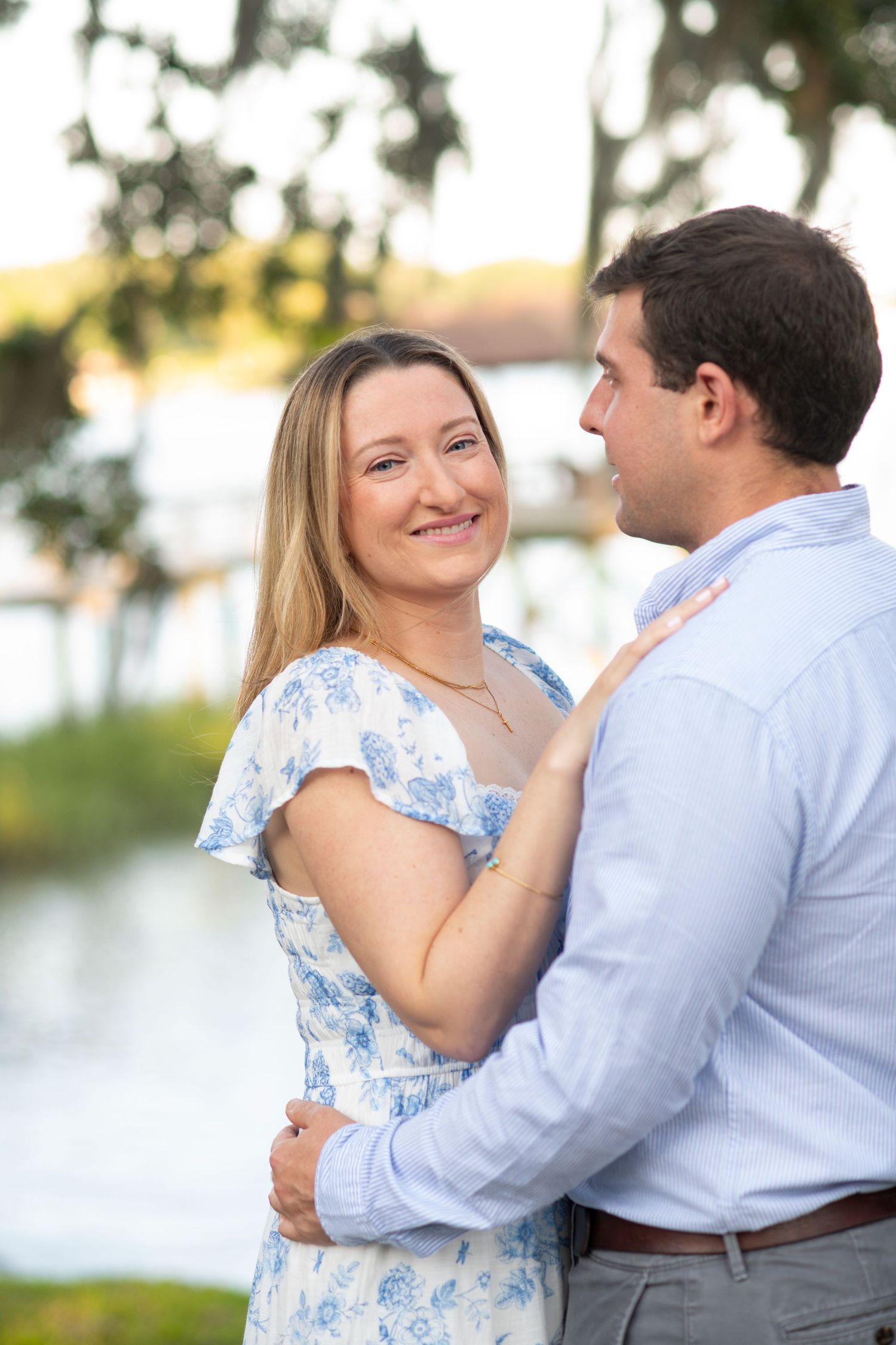 Romantic couple in light blue and white attire share tender moments during outdoor photoshoot by the water.
