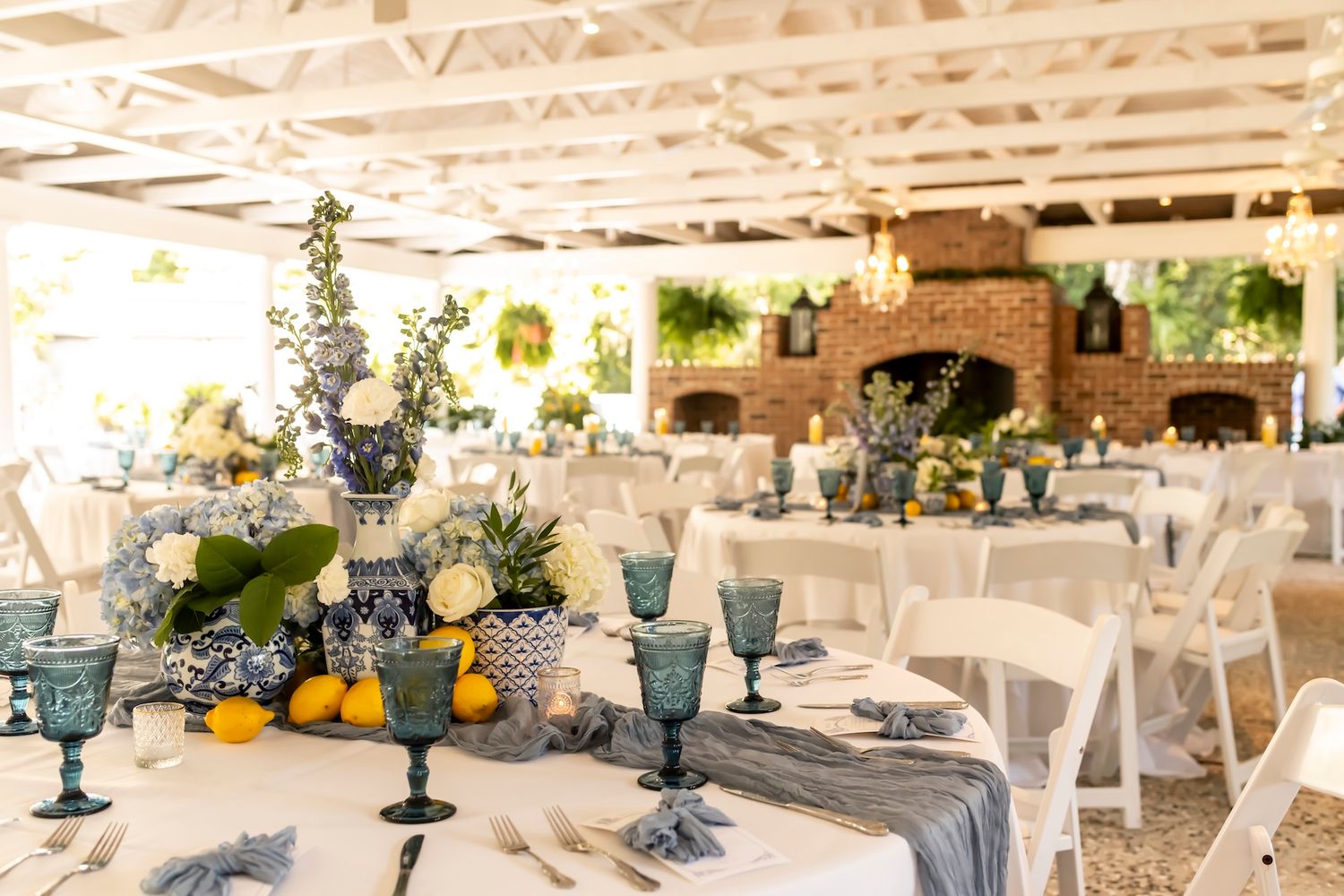 Elegant wedding reception tables with blue glassware, floral centerpieces, lemons, and white chairs in a bright venue.