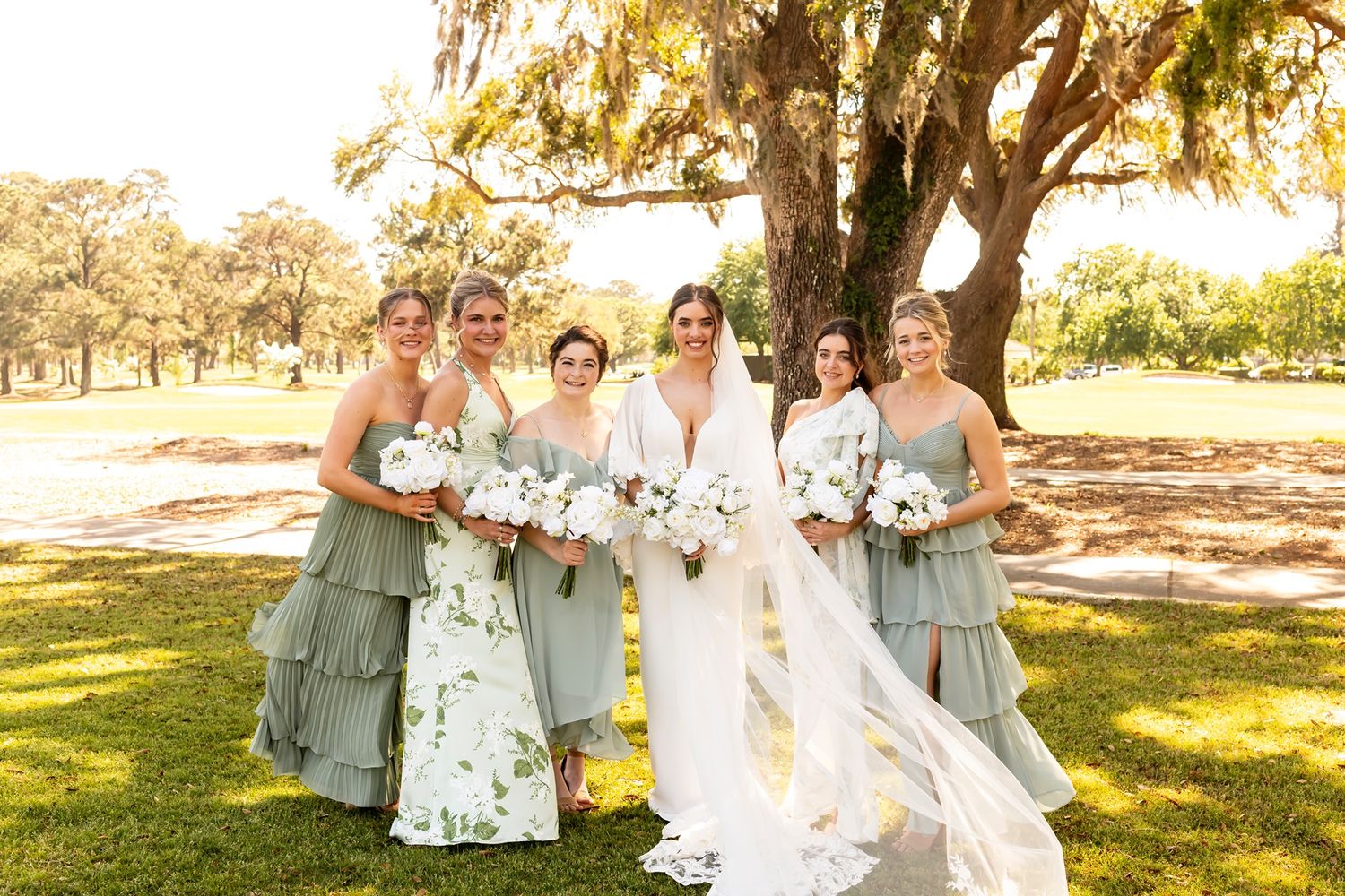 A bridal party wearing gray and white dresses poses together outdoors in a sunlit garden setting.