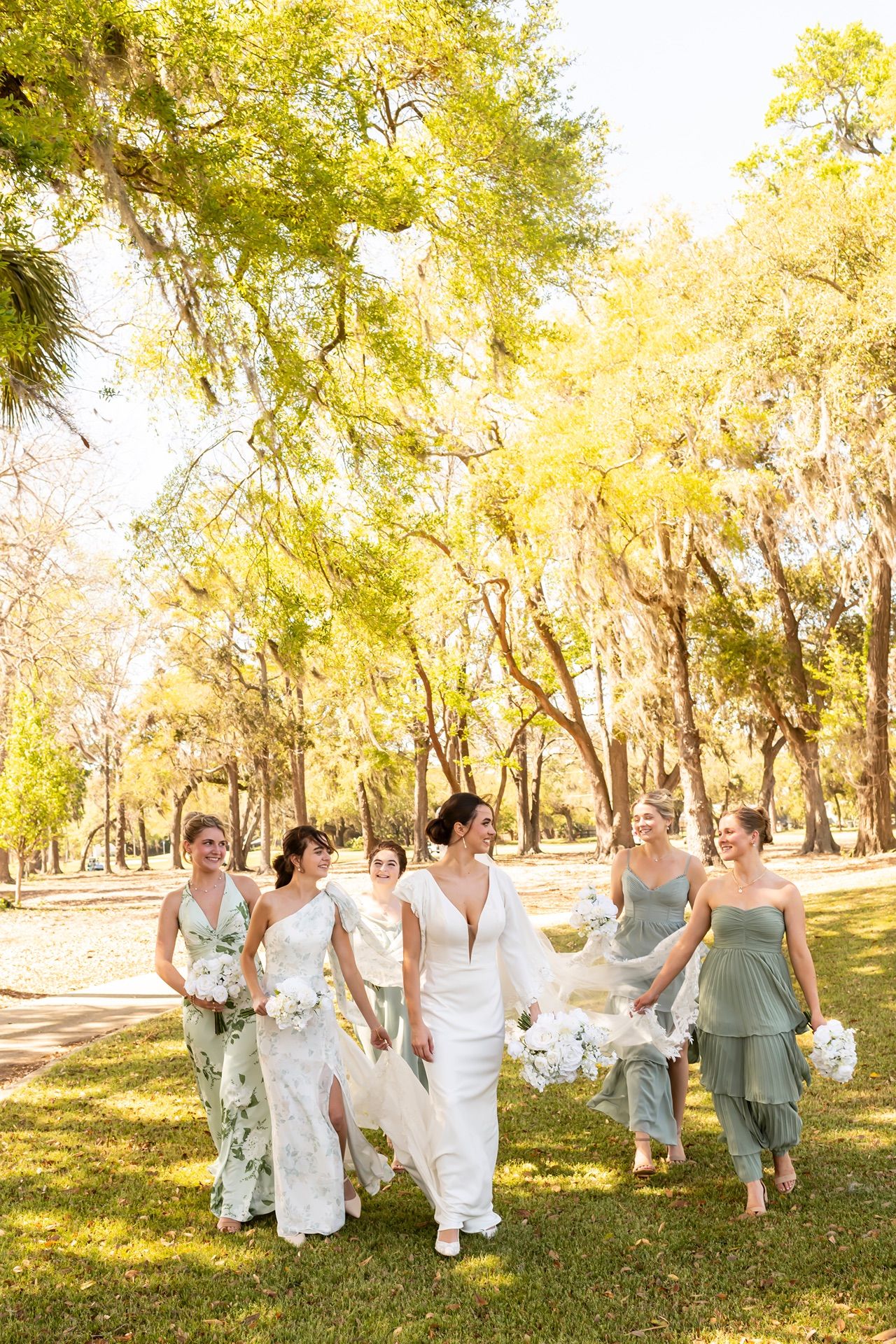 A wedding party dressed in white and sage green runs playfully through a sunlit park with autumn trees.