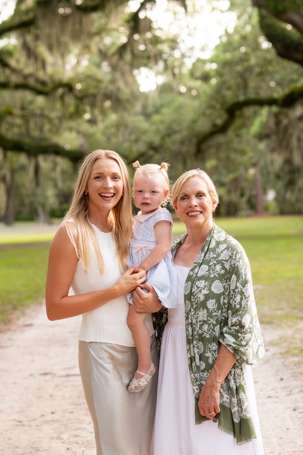 Three generations of women smiling outdoors under oak trees, mother holding toddler girl in a lush green park setting.