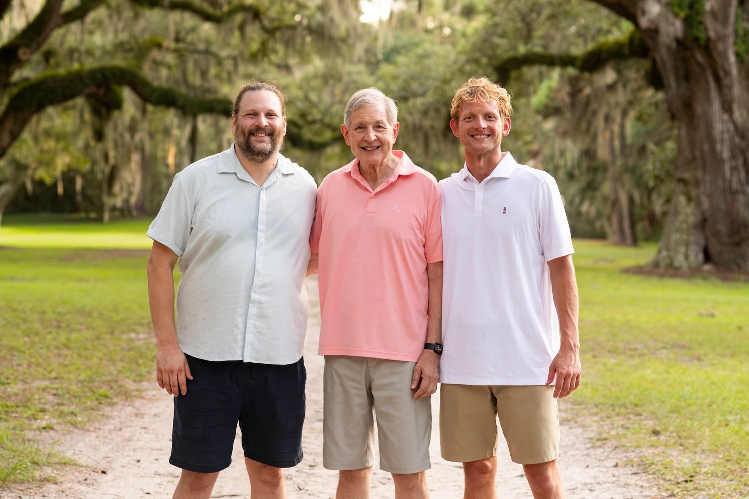 Three smiling men standing together outdoors on a dirt path surrounded by lush green trees.