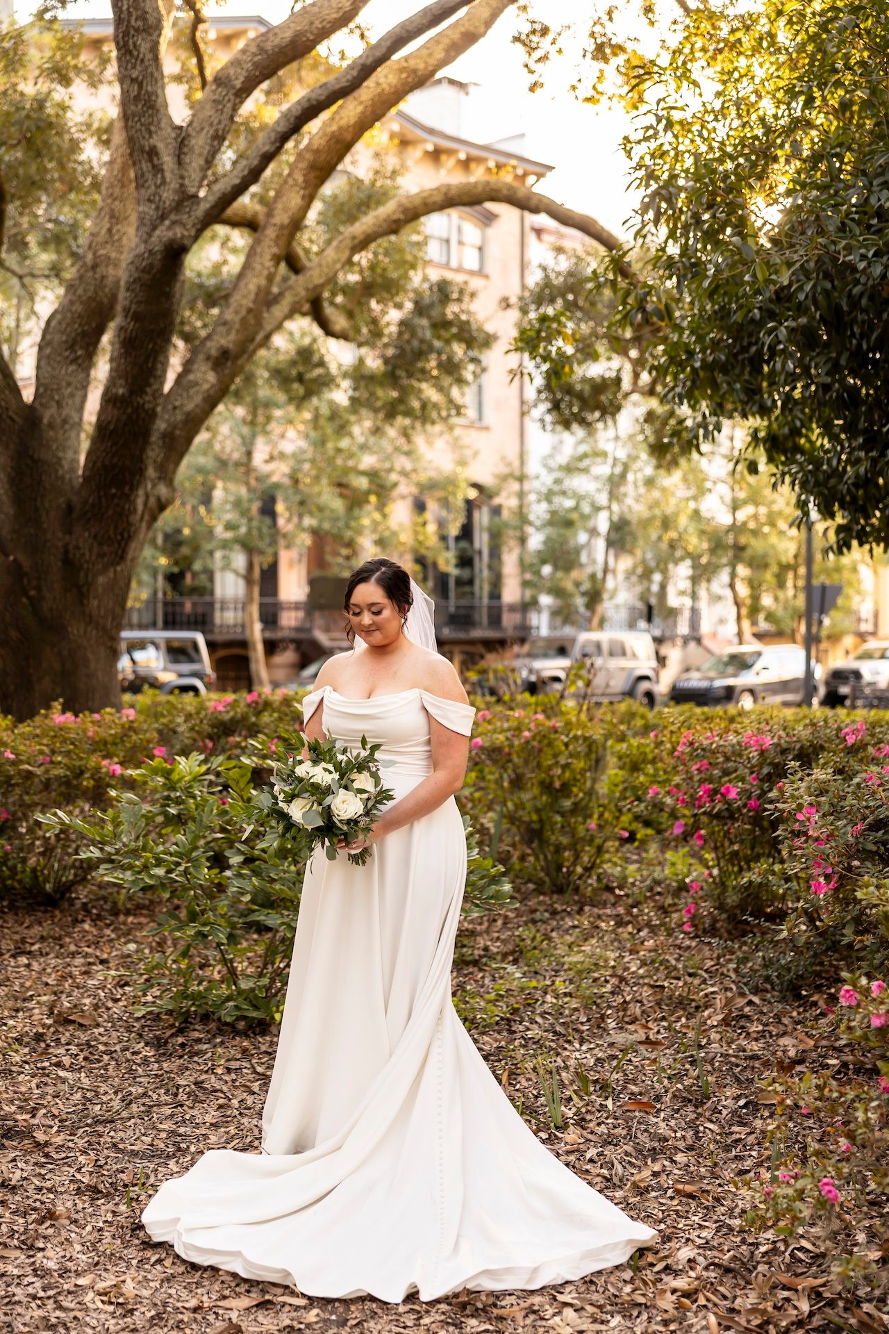 Bride in elegant off-shoulder white gown holding floral bouquet poses outdoors in garden setting during autumn.